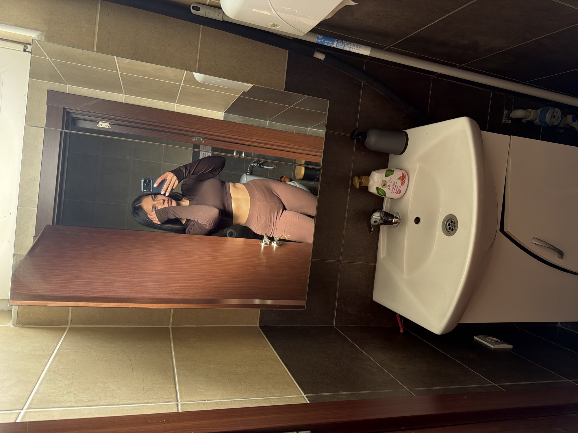 The image depicts a person reflected in a bathroom mirror. The individual, dressed in a brown long-sleeved top and matching leggings, is holding a phone and partially covering their face. The bathroom features brown and beige tiling on the walls and floor. A white sink and vanity unit are visible to the right of the mirror. A bottle of hand soap and a black dispenser are on the sink. The environment appears to be a typical domestic bathroom setting. There is no verifiable information indicating the location in Gabrovo, Bulgaria.
