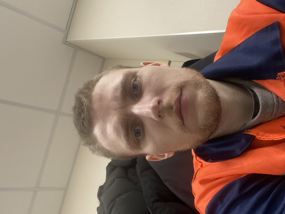 A male individual, appearing to be in his 20s or early 30s, is depicted in a close-up, head-and-shoulders portrait. He has short, light brown hair, blue eyes, and light stubble or a short beard. His expression is neutral, and he is looking directly forward. He wears an orange and navy blue two-tone work garment over a grey t-shirt. The background on the left features a dark, possibly black, puffy jacket or coat. On the right, a light-colored wall or cabinet with a vertical edge is visible. The overhead area reveals a light-colored grid-style suspended ceiling. The scene takes place indoors in the city of Nikol'skoe, Russia, likely within an office or a similar institutional setting.