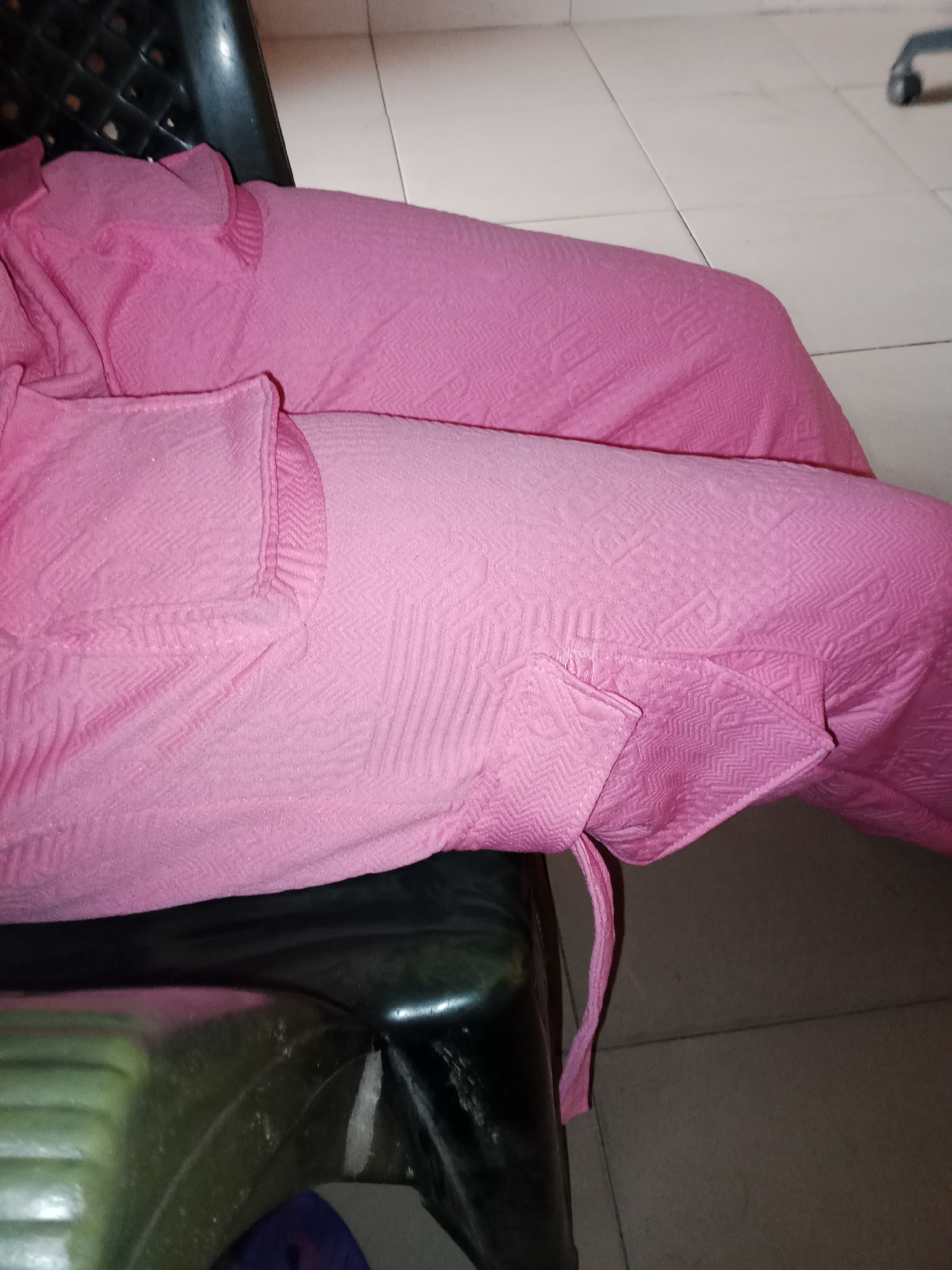 In Jalingo, Nigeria, a close-up shot captures pink bedding draped over a dark plastic chair. The bedding features a textured pattern of small zigzags and vertical lines, with a ruffled edge visible on the left side. A pink tie or strap is hanging down from the bedding on the right. The background consists of light-colored tiled flooring, with a blurred object and its wheel visible on the right edge. The scene appears to be indoors.