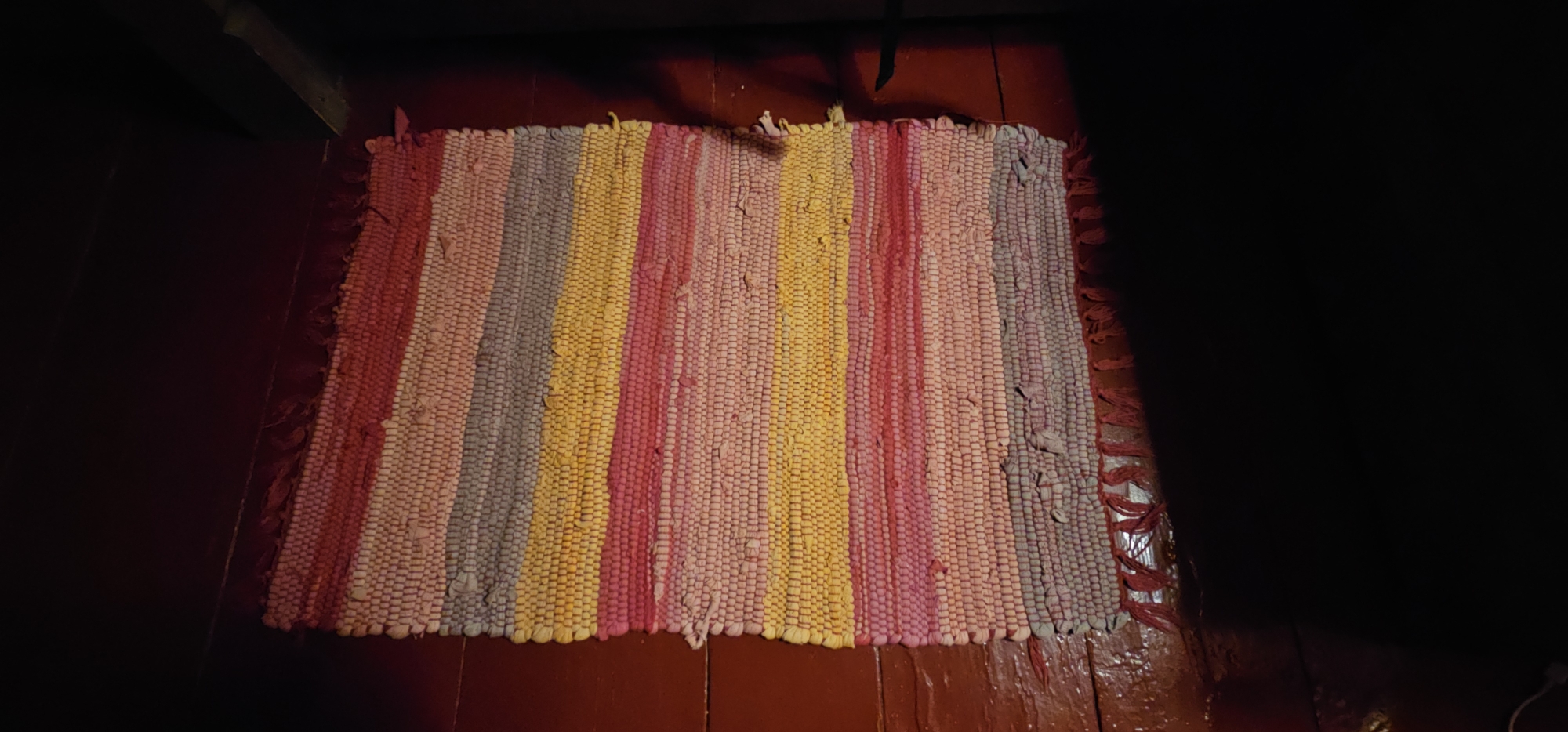 An overhead, slightly angled, medium shot captures a rectangular, handmade rag rug with vertical stripes of various colors, including pink, yellow, light gray, and red, lying on a dark red wooden floor. The rug features a woven texture and frayed fringe on its edges. The lighting is dim, casting shadows across the scene, particularly on the right side, which is almost entirely black. The top left of the image shows a dark wooden surface, possibly a piece of furniture or part of a wall, with a sliver of light visible at the very top edge, suggesting it might be a corner or a gap. The overall impression is of a dimly lit interior space. The location is identified as Plyos, Russia.