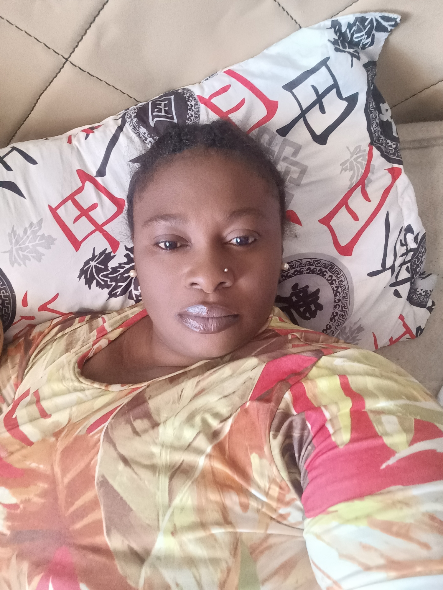 A woman is lying down on a beige quilted surface with a pillow under her head. The pillow is white with red and black Asian-inspired characters and designs. She is wearing a colorful shirt with red, yellow, and brown patterns. She has dark hair pulled back, a nose piercing, and glossy lips. The location is likely indoors and given the prompt is presumed to be Jos, Nigeria, but there are no visual clues to confirm this.