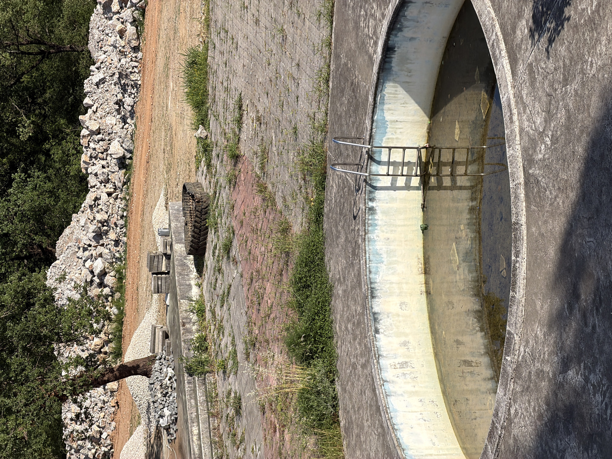 A top-down view shows a circular concrete structure containing water, possibly a retention pond or abandoned swimming pool, in an outdoor area, likely in Bratešići, Montenegro. A metal ladder is fixed inside the concrete ring. The exterior of the circle is paved with interlocking bricks, some overgrown with grass and weeds. Adjacent to the paving is a dirt area containing discarded construction materials like a tractor tire and stacked concrete blocks. Beyond that is a rock pile next to a tree and dense vegetation. The scene is mostly static and contains no identifiable people, interactions, or actions. The overall environment appears to be a rural or semi-rural site, perhaps undergoing or awaiting further construction or development.