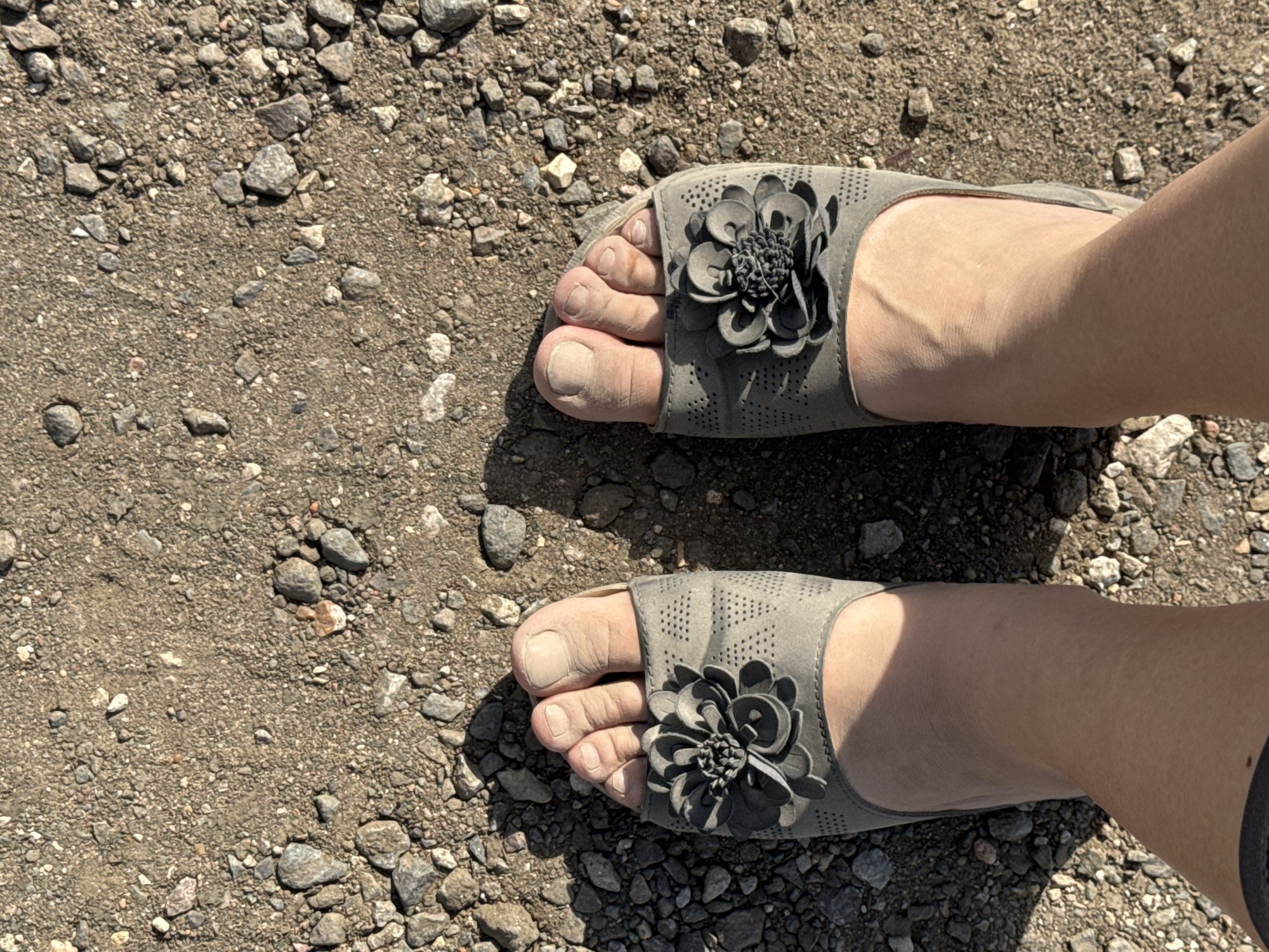 The image shows a close-up, overhead view of two feet wearing grey slide sandals with a flower embellishment. The feet are resting on a surface of dirt and small stones. The lighting suggests it is daytime, likely sunny, as there are shadows cast by the feet and sandals. The setting appears to be outdoors, possibly a gravel path or unpaved ground. The sandals are a light grey color and have perforated detailing and a dark grey, layered flower attached to the strap. The toenails are visible and appear unpolished. The skin on the feet and ankles shows some texture and veins. There is no visible text in the image. The context provided places this scene in Markovo, Russia.