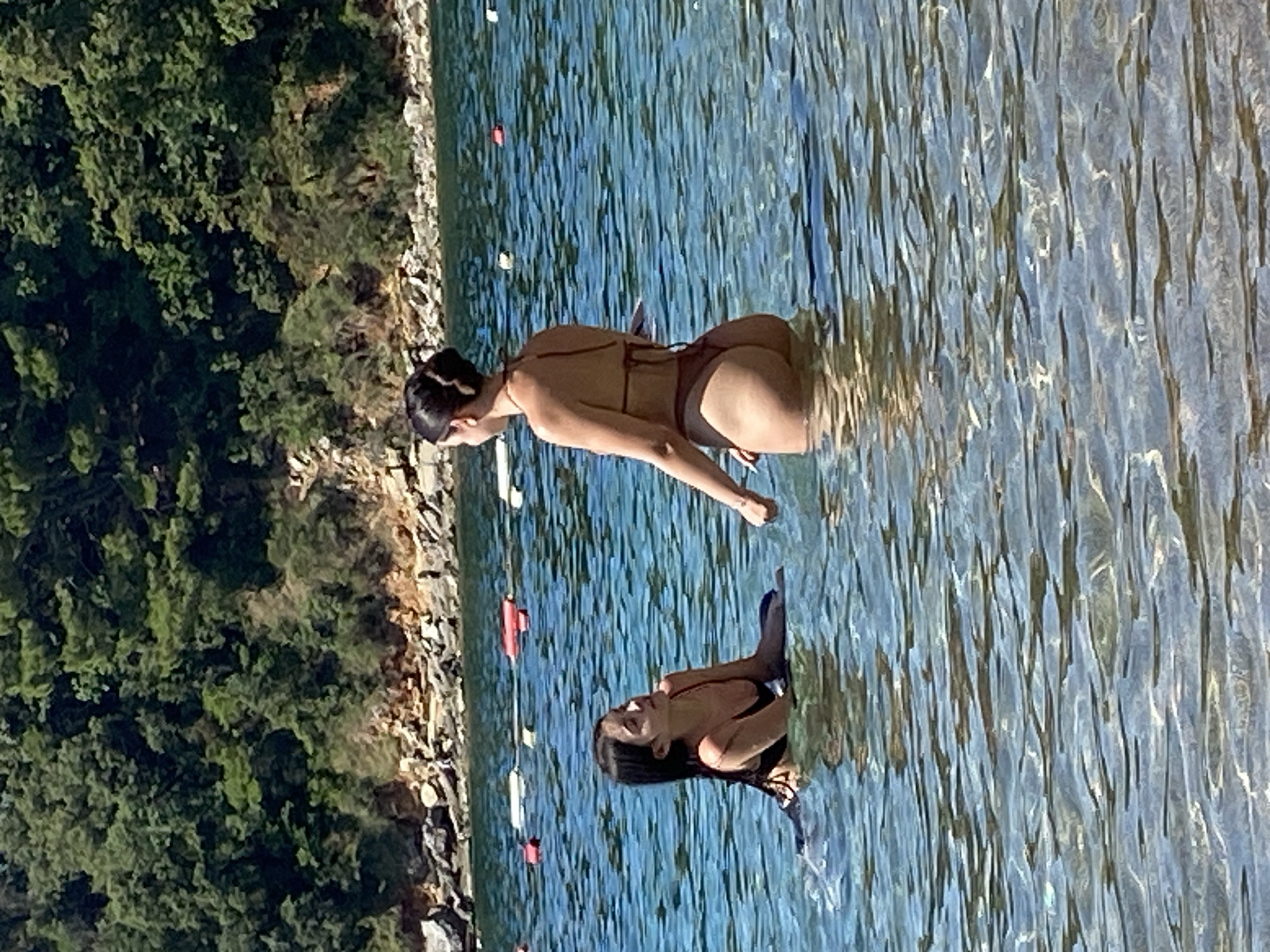 In Budva, Montenegro, two women are standing in clear, shallow, turquoise water near a rocky, tree-lined shore. The woman on the left, with long dark hair, is wearing a black bikini and appears to be lying down in the water, looking towards the woman on the right. The woman on the right has her dark hair pulled up in a bun and is wearing a brown bikini. She is facing away from the camera, looking towards the woman on the left. The water has visible ripples and sunlight reflects off the surface. In the background, red and white buoys are floating in the water, marking a designated swimming area. The shore is covered with dense green trees and scattered rocks.