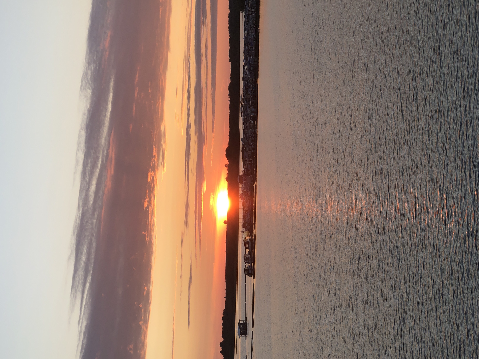The image captures a sunset over a body of water, likely a river or lake, in Solikamsk, Russia. The sun, a bright orb, is low on the horizon, casting a warm, orange glow across the sky and reflecting on the rippling water. The sky is filled with dramatic clouds, streaked with shades of orange, pink, and purple, creating a vibrant and atmospheric backdrop for the sunset.
Along the shoreline, a dark line of trees and vegetation is visible, separating the water from the sky. A small settlement or marina appears to be nestled along the coast, with several small boats moored near the shore. One structure, possibly a small boathouse or shed, is situated directly on the water's edge.
The overall scene evokes a sense of tranquility and natural beauty. The time of day is clearly sunset, and the weather appears calm, with the water showing only gentle ripples. The location in Solikamsk, Russia, suggests a possibly northern landscape. No text is visible in the image.
