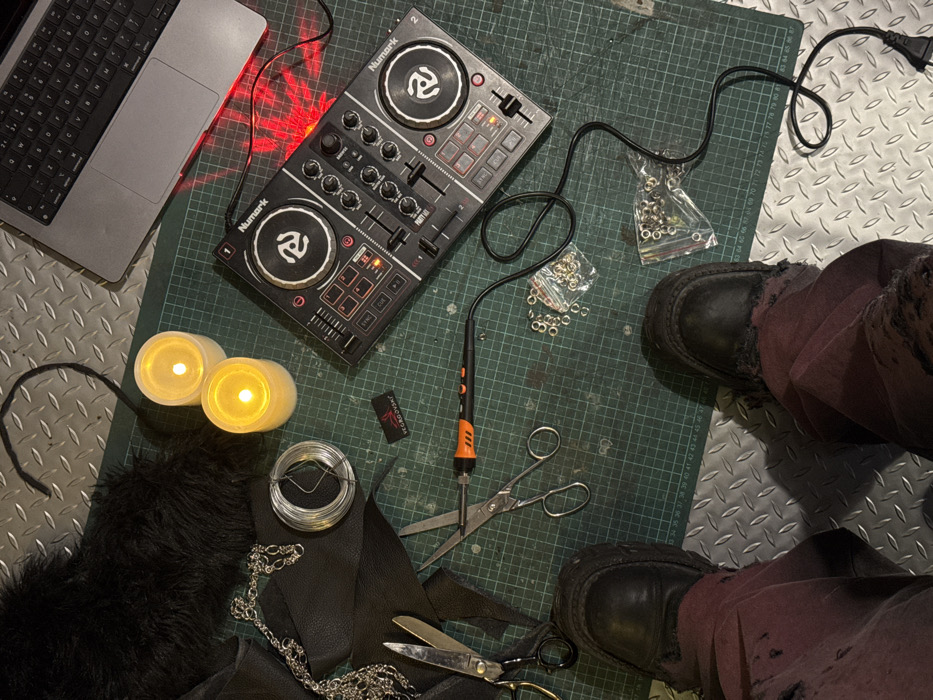 The scene depicts an indoor workspace in Sofia, Bulgaria, viewed from a top-down perspective. A person's lower legs are visible at the bottom of the frame, clad in dark, distressed fabric trousers and black, chunky platform shoes, positioned on a metallic diamond-plate floor.
The primary work surface is a large, dark green self-healing cutting mat with a white grid and measurement markings. Centrally located on the mat is a black Numark Party Mix DJ controller, powered on and emitting red LED light. A silver-colored laptop is open to the upper right of the controller. Two white, cylindrical LED candles emitting a warm yellow light are situated on the left side of the mat.
Various crafting materials are present: a spool of thin, silver-colored wire, several irregularly cut pieces of black leather or faux leather fabric, a silver-colored metal chain, and a piece of black faux fur. A small black fabric tag labeled "VEGROVSKY" is visible near the candles. Crafting tools include two pairs of silver-colored metal scissors and an orange and black soldering iron or heat pen with a black power cable. Two clear plastic bags containing numerous silver-colored metal grommets or eyelets are also on the cutting mat. The arrangement suggests an ongoing crafting project alongside music equipment.