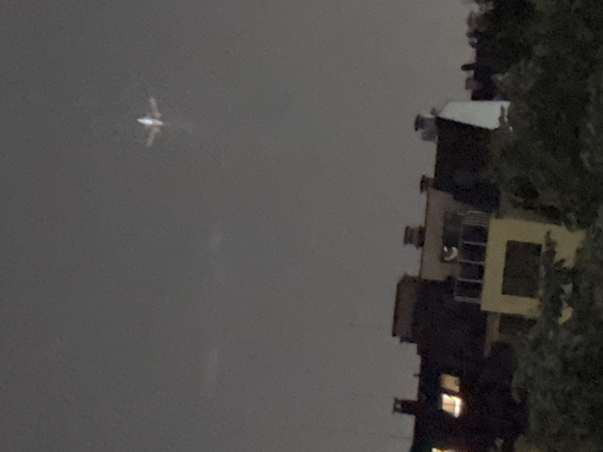 The image captures a nighttime urban scene, with a brightly lit airplane prominently featured against a dark sky. The airplane, rendered somewhat blurry due to motion or distance, appears white and is seen from below, showcasing its fuselage and wings extended horizontally. It occupies the left half of the frame, suggesting it is flying overhead.
On the right side of the image, a multi-story building is partially visible, appearing dark with some windows illuminated, indicating residential activity. The architecture suggests a common urban design. A dense, dark tree obstructs part of the building and is also visible on the right. The overall setting implies a typical evening in a city, with air traffic overhead. The sky is dark and featureless, characteristic of night. There is no visible text in the image. The scene is consistent with an urban environment in Sofia, Bulgaria.