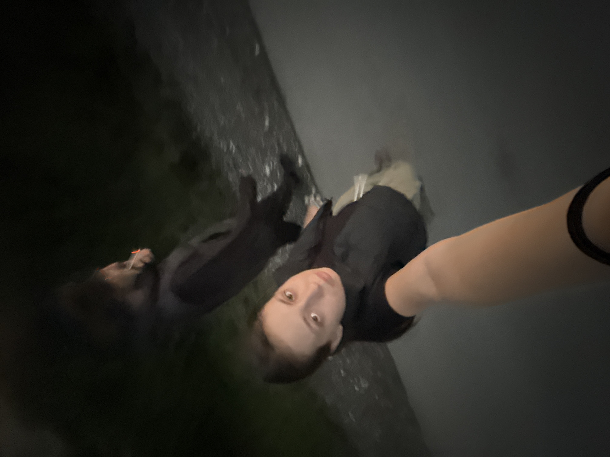 In the city of Radishevo, Bulgaria, a selfie is taken at night by a young woman wearing a black t-shirt and khaki pants. She is standing in what appears to be shallow water, possibly a puddle or a wet road, as evidenced by the reflections. Her left arm, with a black band on her wrist, is extended towards the camera. Behind her, another person, partially obscured and out of focus, is visible, also appearing to be outdoors in the dark. The background is predominantly dark with some faint hints of green foliage, suggesting an outdoor environment at night. The overall lighting is dim, indicating the absence of strong artificial light sources.