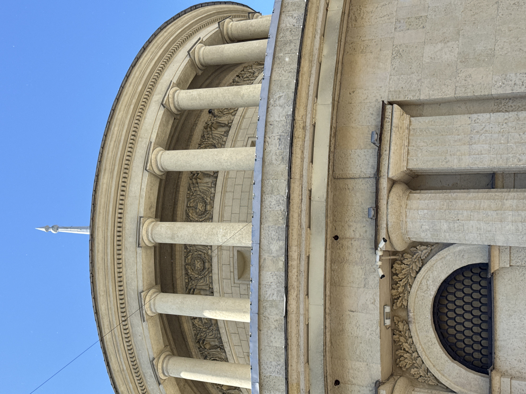 This is a low-angle, exterior, eye-level shot of a large, rounded, light-colored stone building with classical architectural elements against a clear blue sky. The building features a prominent rotunda-like structure with a series of large, fluted columns supporting an upper level. Above the columns, there is a decorative frieze with relief carvings and an ornate cornice. The uppermost part of the building visible shows a curved roofline and a spire-like finial.
Several security cameras are mounted on the facade of the building. Below the columns, there is an arched window with a dark, decorative metal grate. The stonework of the building is detailed, with visible mortar lines and some signs of weathering. A single black wire runs diagonally across the upper portion of the image against the blue sky, possibly a utility line. The overall impression is of a grand, historical public building. This building is the Kazan Cathedral, located on Nevsky Prospekt in Saint Petersburg, Russia.