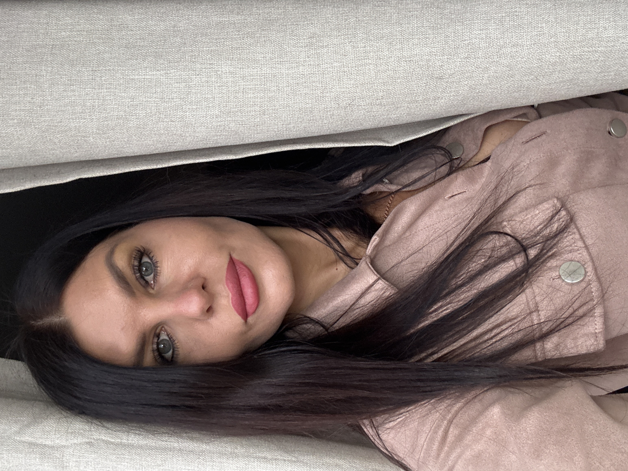 The image displays a young woman with long, dark hair and striking blue-grey eyes, lying on her side and looking directly at the camera. She appears to be nestled between or under light-colored, textured fabric surfaces, possibly sofa cushions or bedding, with a similar fabric surface positioned above her, creating a narrow opening from which she peeks out.
Her face is framed by her dark hair, and she has visible makeup, including bright pink lipstick and mascara that accentuates her eyes. She is dressed in a light pink or dusty rose-colored jacket, featuring silver buttons on the front, and a thin gold chain is visible near her neck. The area behind her head is dark, contrasting with the lighter fabric surrounding her.
The activity captured is a direct gaze at the viewer, suggesting a posed portrait. Her expression is calm and direct, conveying a subtle allure. The setting appears to be an intimate, indoor, domestic environment, possibly within a home in Chaikovsky, Russia. There is no visible text in the image.