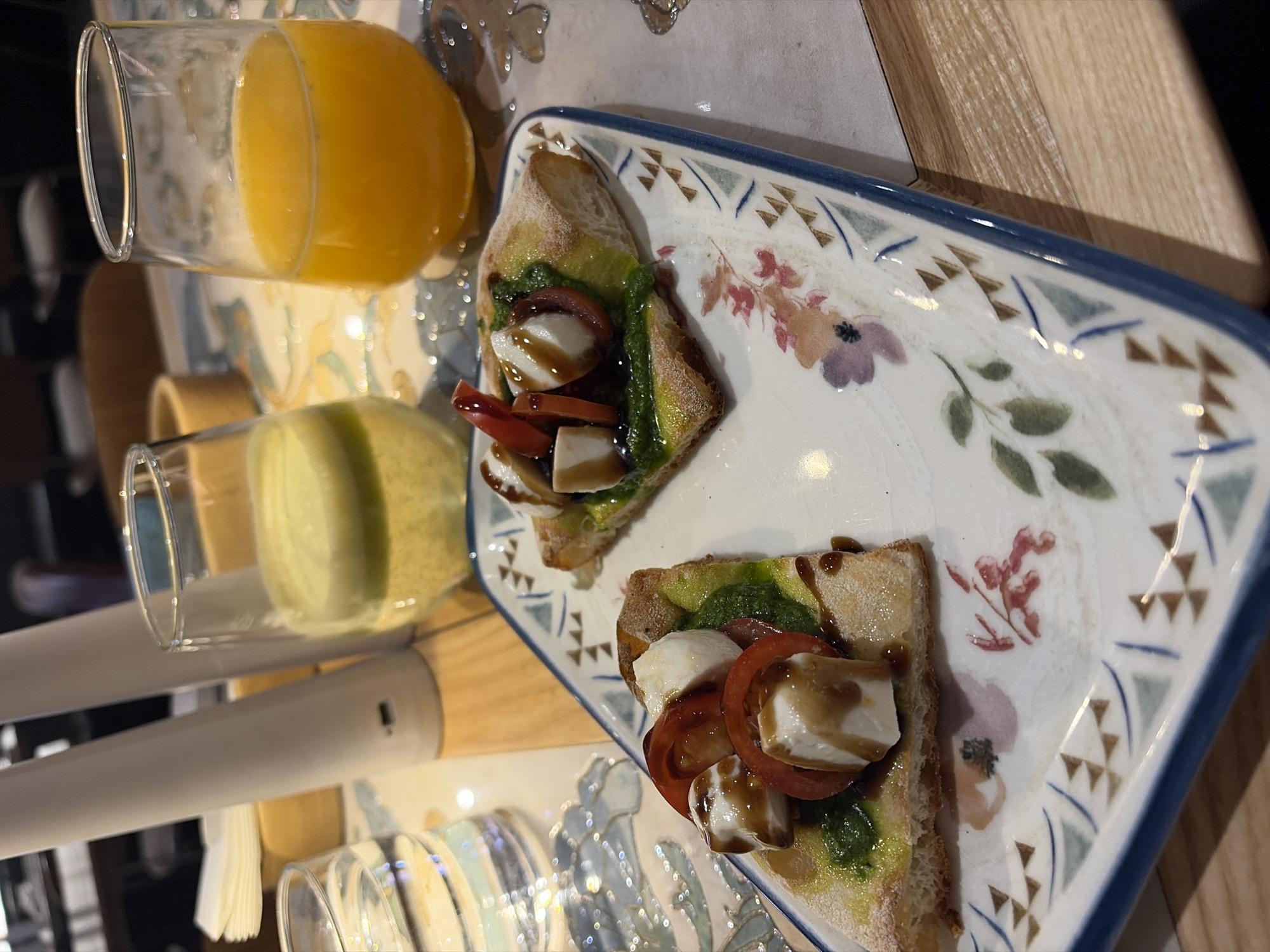 The image depicts a close-up, elevated view of a dining scene, consistent with an indoor restaurant or cafe setting in Perm, Russia. The specific location within Perm is not identifiable from the visual evidence.
The central focus is a rectangular white ceramic plate with a decorative blue rim, featuring a floral and geometric pattern. On this plate are two individual portions of bread, likely focaccia or flatbread. Each portion is topped with a layer of green pesto, two slices of red tomato, several irregular cubes of white mozzarella, and a drizzle of dark balsamic glaze.
Adjacent to the plate, three clear, stemless drinking glasses are present. From left to right: one glass contains clear water, another contains a pale yellow-green liquid, and the third contains an orange liquid.
The table surface beneath the items features a central patterned inlay with blue, white, and beige motifs, surrounded by a light-toned wooden surface. In the blurred background, two tall, white, conical objects are visible, one of which displays a USB port. A stack of white napkins is also partially visible behind these objects. Further background elements are indistinct, suggesting furniture and the general ambiance of a dining establishment. No individuals are discernible within the frame.