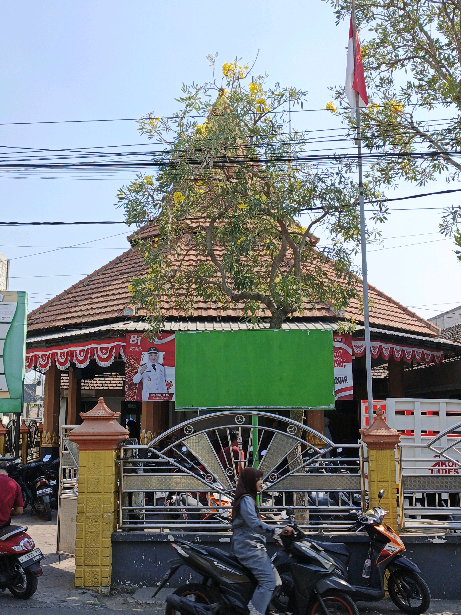 In Ketegan, Indonesia, a sunny day is depicted with a building featuring a prominent multi-tiered, tiled roof topped by a single tree with yellow flowers. A flagpole with a red and white Indonesian flag stands to the right. Several overhead wires crisscross the sky. A green, rectangular sign is mounted on the building's facade, obscuring part of a banner with a portrait of a woman in uniform and the number "81". Red and white decorative bunting adorns the front of the building. A metal gate with an ornate circular design separates the street from the building's entrance. Various motorcycles are parked or being ridden in front of the gate, including a woman wearing a hijab riding a black scooter. A red motorcycle is parked to the left, and a large white truck with red accents is visible in the background. The environment appears to be a commercial or public area with a sidewalk and street.