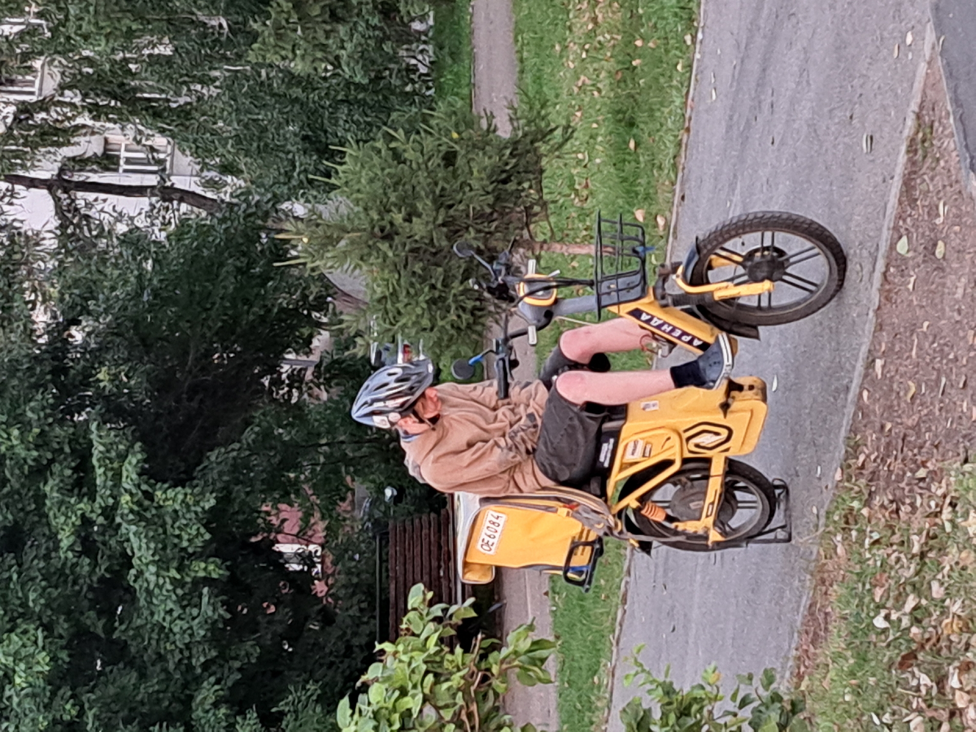 A young male wearing a helmet and a tan hoodie is sitting on a yellow electric scooter with a large yellow delivery box on the back. The scooter has the license plate OE6084. The scooter is parked on a paved path next to a grassy area with fallen leaves. In the background, there are trees and a building. The scene appears to be a park or residential area. The scooter has the word "АРЕНДА" (ARENDA, meaning "rent" in Russian) printed on the front.