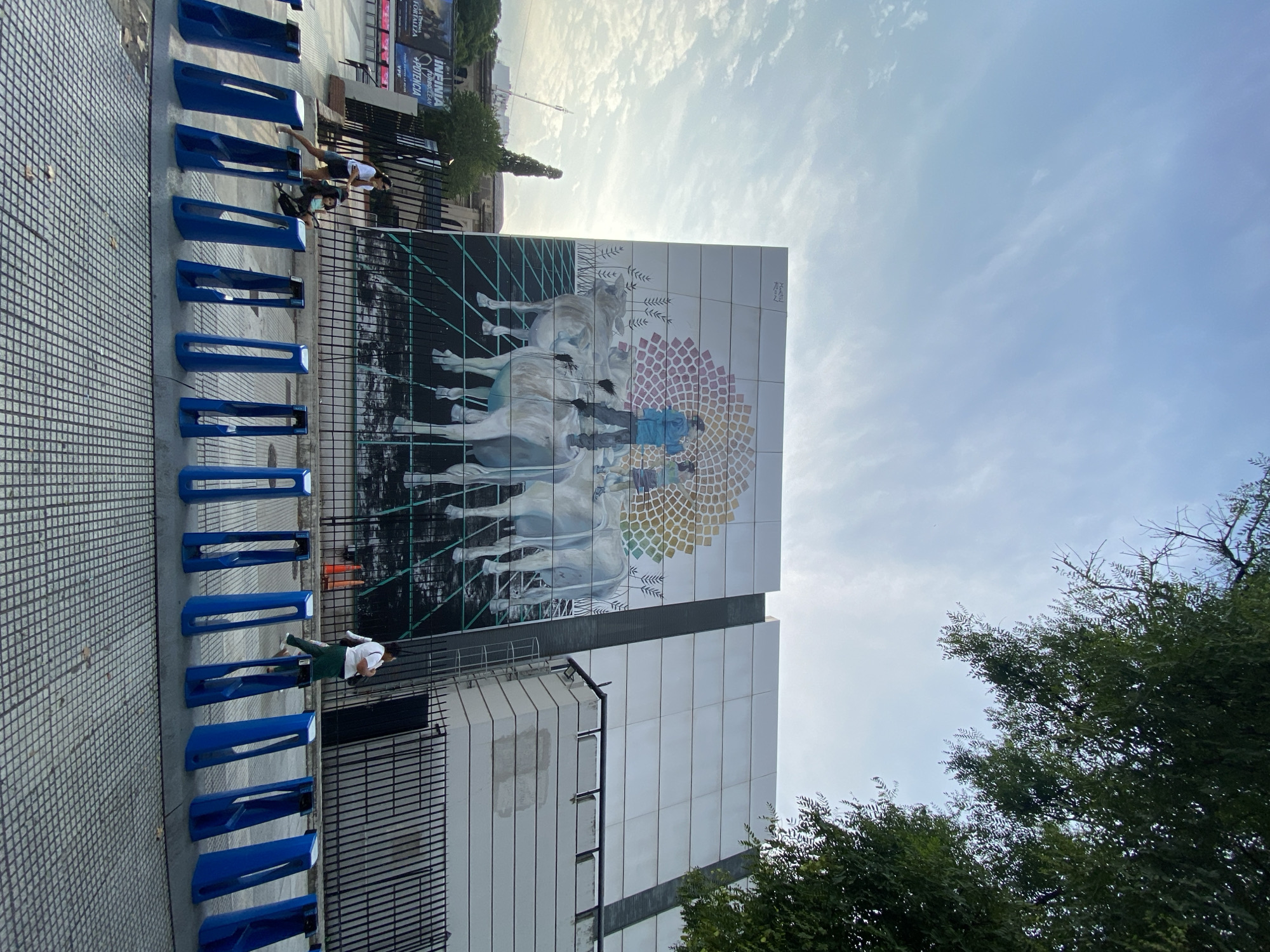 The image depicts a modern building in Buenos Aires, Argentina, adorned with a large mural featuring horses and human figures, rendered in a mix of realistic and stylized techniques. The sky is partly cloudy. A row of blue, modern-designed bicycle racks lines the sidewalk adjacent to the building. Two individuals are standing near the bike racks. Vegetation, specifically a tree, is visible in the lower right corner of the frame. The sidewalk is paved with a square-patterned material. In the background, there is a billboard.