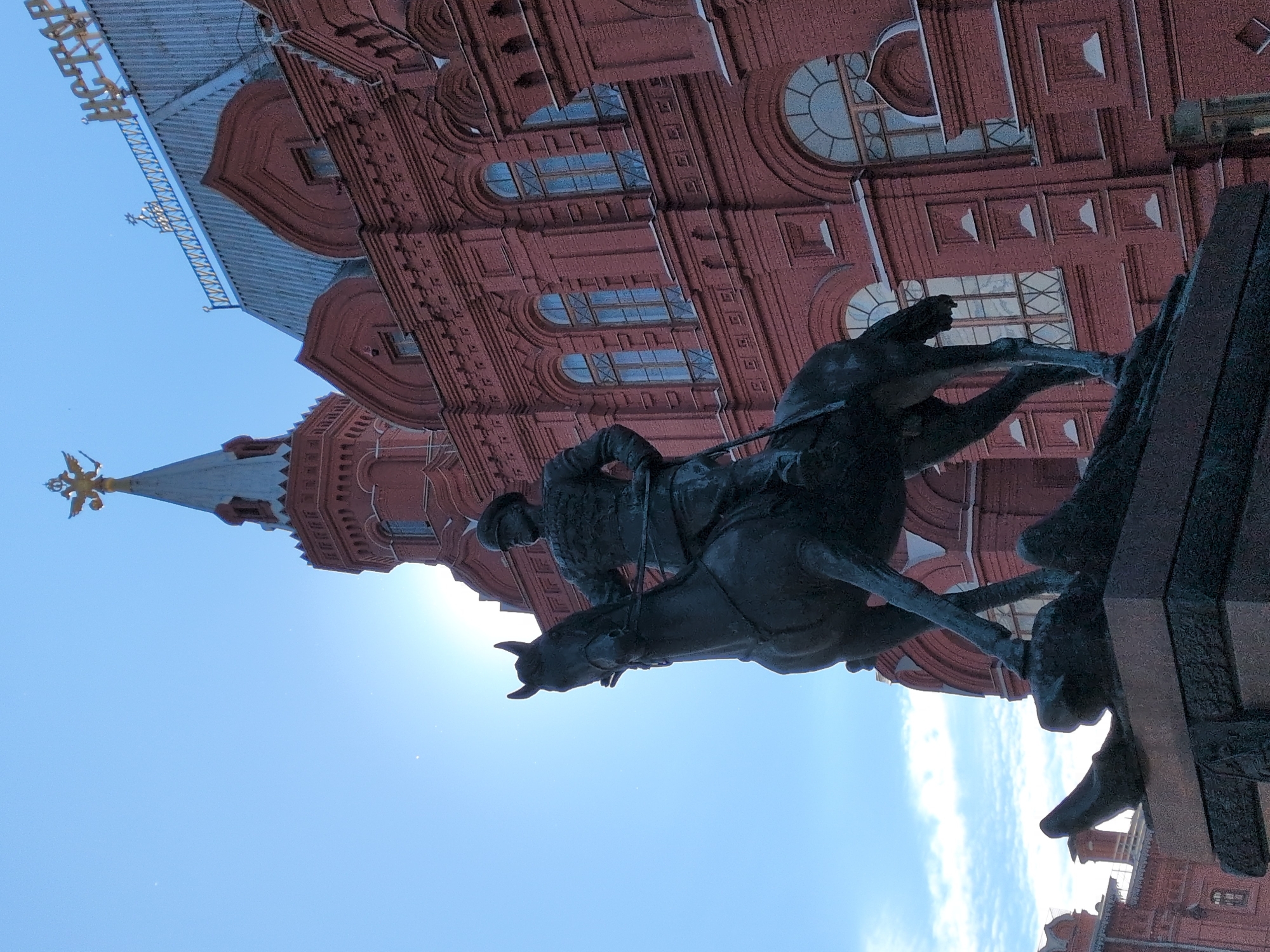 The image captures a monumental bronze equestrian statue set against a detailed red brick building under a bright, clear blue sky. The photo is rotated 90 degrees counter-clockwise.
In the foreground, a large bronze statue depicts a man in what appears to be historical military attire, including a helmet, mounted on a rearing horse. The man holds the reins, and the horse is depicted in a dynamic, powerful pose. To the right, another section of a bronze monument is visible, likely part of a larger sculptural group or base.
Behind the statue, a grand red brick building dominates the upper portion of the frame. Its facade is richly decorated with elaborate architectural details, including arched and rectangular windows, some with intricate grilles, and various decorative elements like cornices and pilasters. A prominent tower on the left side of the building is topped with a distinctive golden finial, which appears to be a double-headed eagle.
The scene is bathed in bright sunlight, with a visible sun flare in the upper right corner of the rotated image, indicating a sunny day. The sky is a vibrant, cloudless blue.
On the roofline of the building, near the top left of the rotated image, some light-colored Cyrillic text is visible, partially spelling out "ИСТОРИЧЕСКИЙ" (Istoricheskiy), which translates to "Historical".