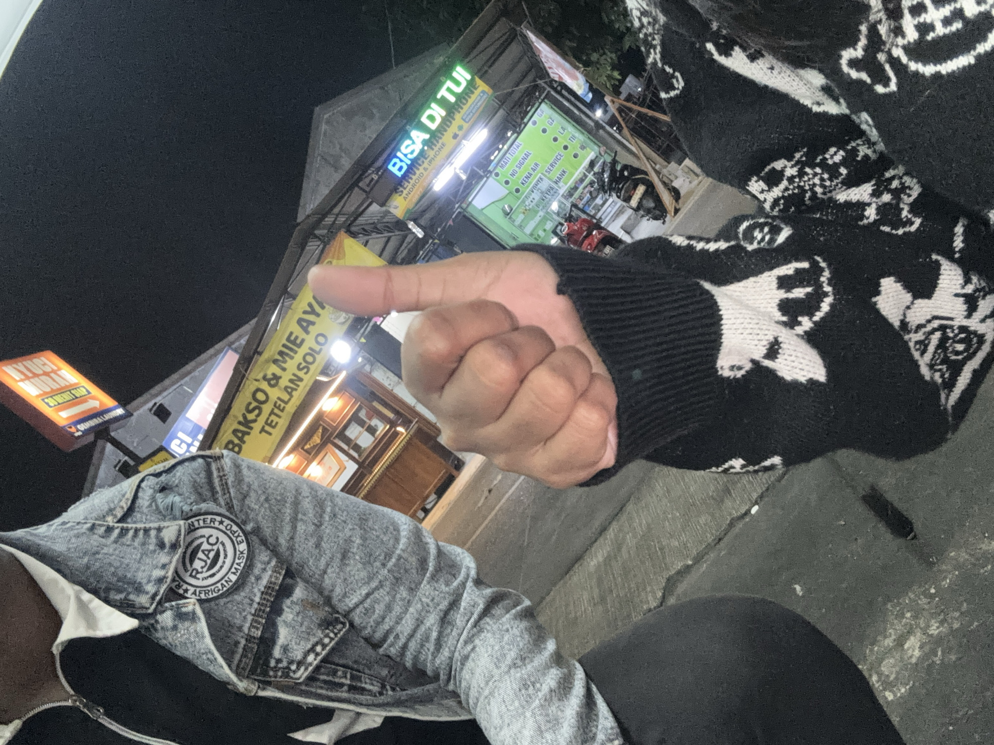 A close-up, low-angle shot shows two individuals outdoors at night. The person on the left wears a light blue denim jacket with a patch on the left breast that reads "RJAC AFRICAN MASK EXPO". They have a white collared shirt underneath. The person on the right has their right hand raised in a thumbs-up gesture, with their left hand visible on their chest. They wear a black sweater with a white skull pattern. In the background, there are several neon signs, including one that says "BAKSO & MIE AYAM TETELAN SOLO" and another in blue and green that reads "BISA DI TUI SERVICE HANDPHONE ANDROID & IPHONE". There is also a wooden display case with framed items and a motorcycle parked in front of a building. The ground appears to be a concrete sidewalk or road. The lighting suggests an urban commercial area.