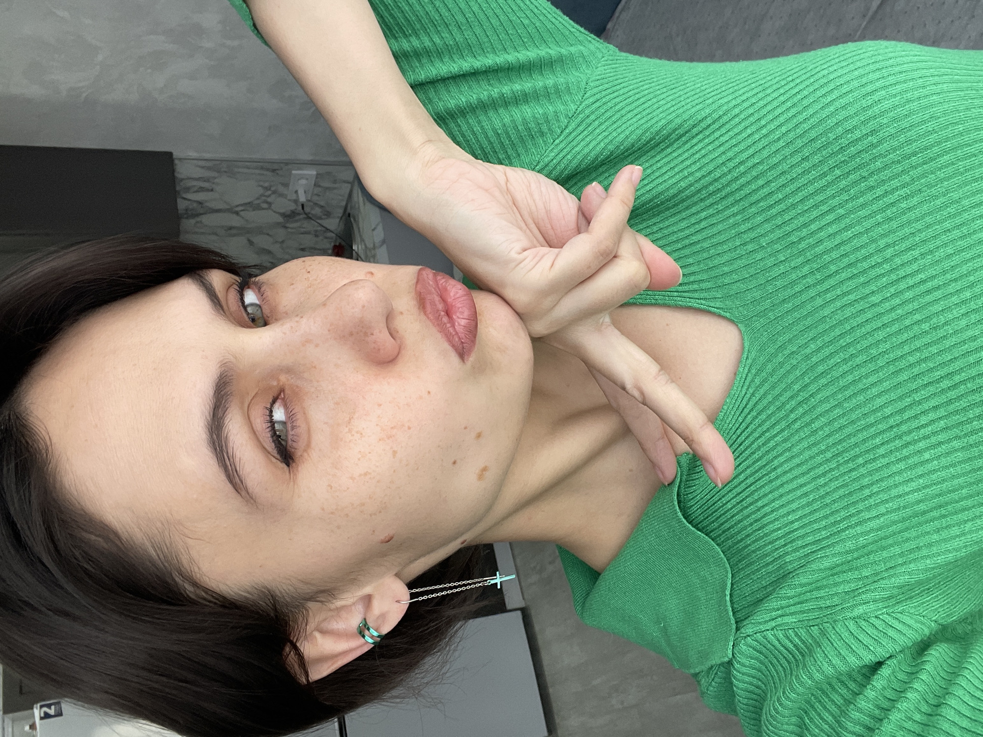 A young woman with dark hair and fair skin poses for a close-up portrait. She has light green eyes and freckles. She is wearing a green ribbed top. Her left hand is near her face, with the fingers curved and her chin resting in her hand. She has a silver chain earring with a cross at the end and a green earring hoop. The background includes a gray wall and the edge of a black cabinet or shelf. No landmarks associated with Izhevsk, Russia are apparent.