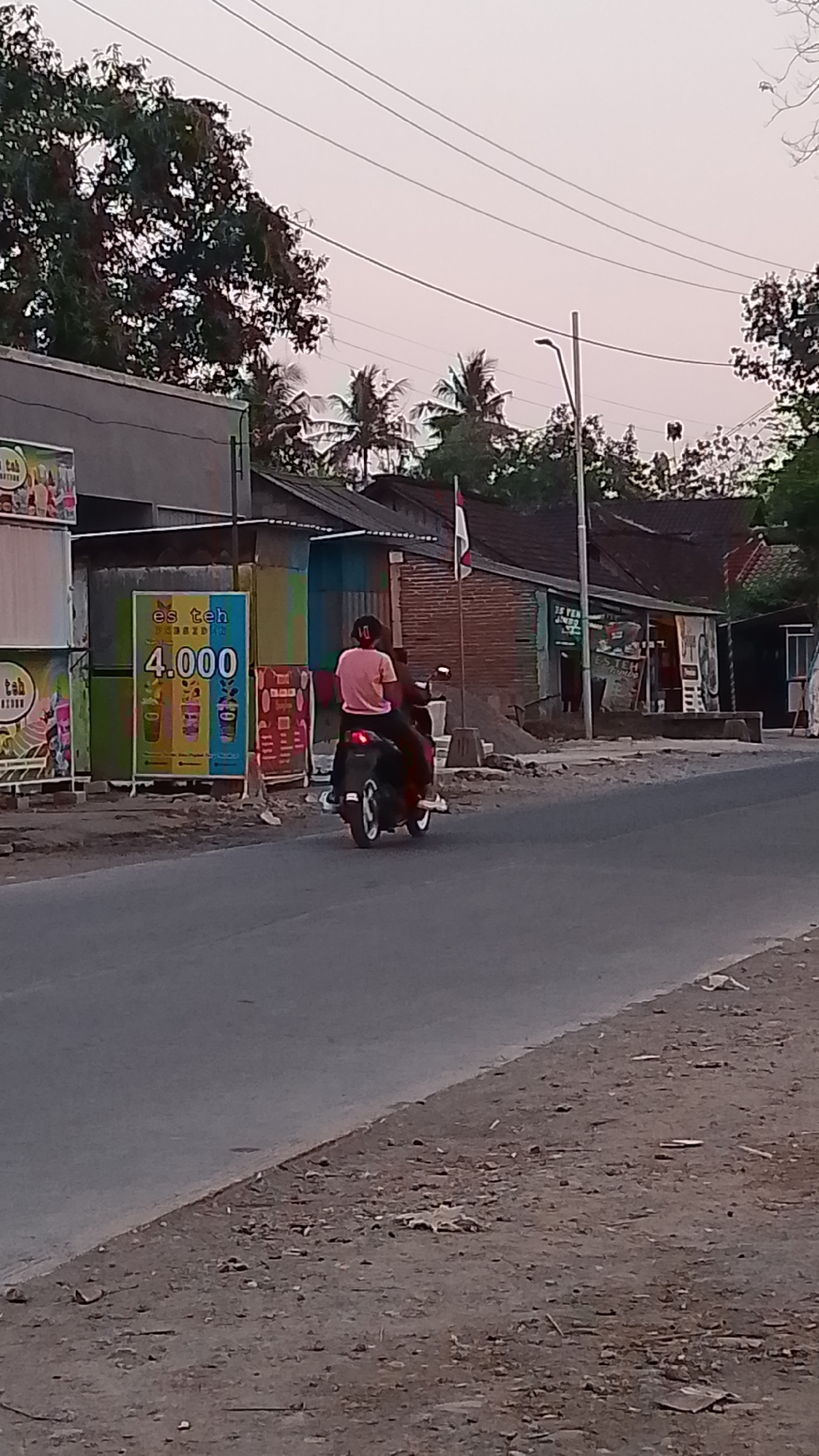 In Karanglangu, Indonesia, a person wearing a pink shirt and dark pants rides a dark-colored scooter on a paved road. They are facing away from the camera. To the left of the road, a tall, colorful billboard displays text and images, including "es teh" and the price "4.000". A light pole with an attached light fixture stands to the right of the billboard, further down the road. Beyond the road, there are buildings with brick and metal facades, and trees with dense foliage. The sky above is overcast and a pale pinkish hue, suggesting it may be dawn or dusk. The ground beside the road is a mixture of dirt, gravel, and debris.