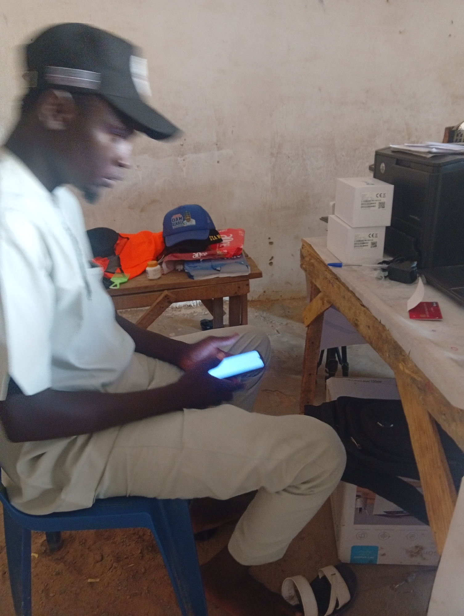 In Mallam Sidi, Nigeria, a young man wearing a white shirt, light-colored pants, and a black baseball cap is seated on a blue plastic chair. He is looking down and interacting with a brightly lit blue smartphone held in his hands. To his right, a wooden table is cluttered with various items, including a blue and white hat with a picture of a man, an orange garment, a small white bottle, and what appear to be stacks of clothes. Further to the right, another wooden table holds a black printer, several white boxes with "CE" markings, a red object that resembles a credit card, and a blue pen. The environment appears to be an indoor space with a light-colored, textured wall.
