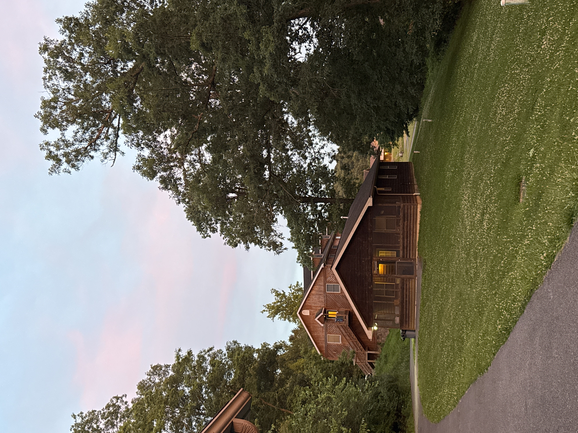 The image shows a scene at dusk in Washington Township, USA, featuring several wooden buildings nestled among lush green trees. A paved path curves through the foreground, leading towards a small, dark brown cabin with a screened-in porch. The cabin has interior lights on, casting a warm glow. Behind this cabin, a larger, multi-story wooden building with a prominent set of outdoor stairs is visible. This building also has lights on, suggesting occupancy. The sky above is a soft blend of pale blue and pink hues, indicative of twilight. The surrounding environment is predominantly foliage, with large trees dominating the right side of the frame and a dense growth of smaller trees and bushes on the left. The grass in the middle ground is short and dotted with small white flowers. The overall impression is of a tranquil, rustic setting, likely a retreat or lodging facility.