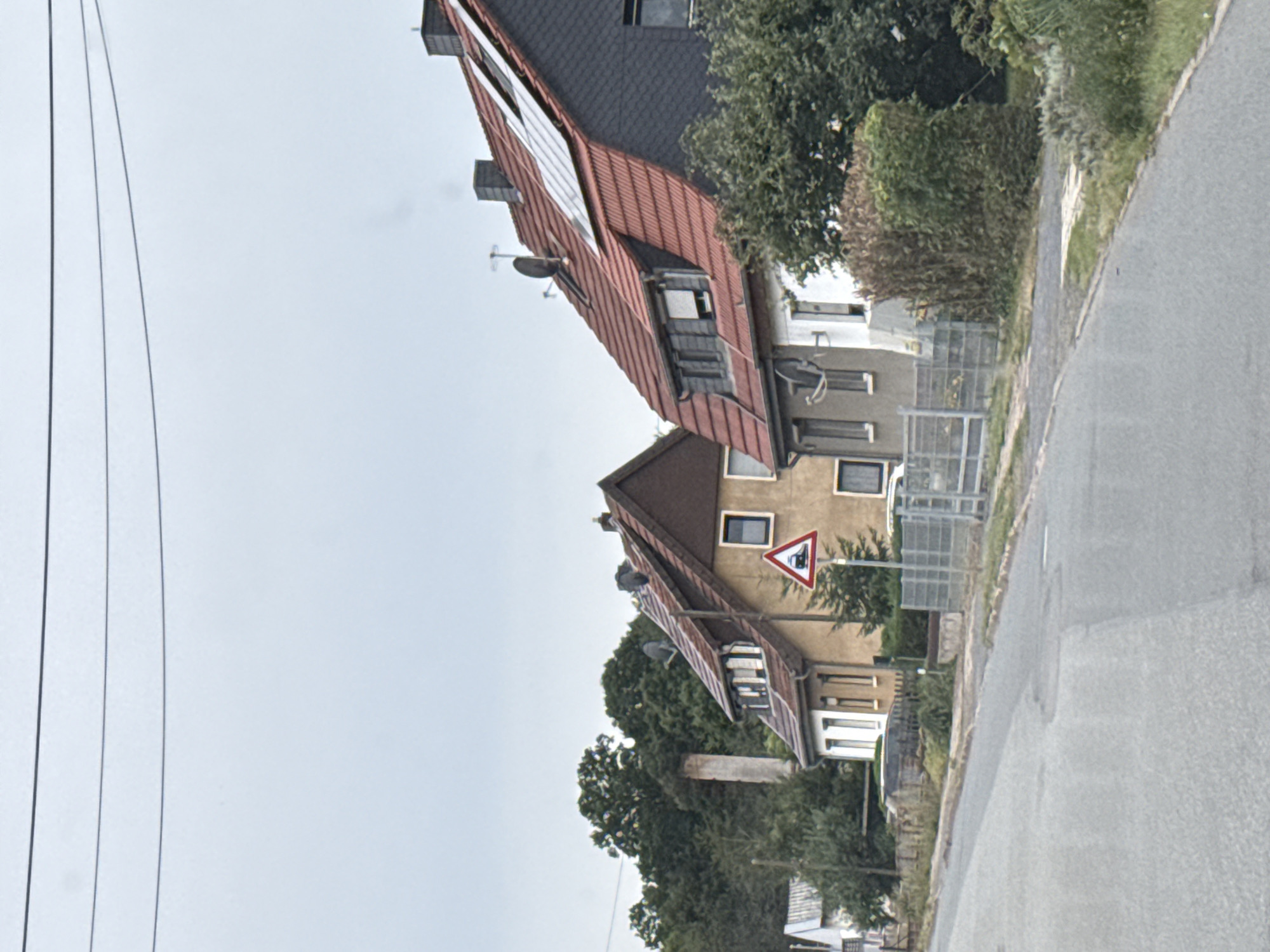 The image depicts a street scene in Ellrich, Germany, under an overcast sky. On the right side of the road are several houses with red tiled roofs. One house, in the foreground, is light brown with white-trimmed windows and a dark gray slate-tiled section on its upper right. It has solar panels installed on the roof. Another house to its left is also light brown with a peaked roof and white-trimmed windows. A red triangular traffic sign with a depiction of a train is visible on a post in front of this house. Further left, a white house with a gray roof is partially visible.

In the background, a tall, narrow, light-colored structure, possibly a chimney or an old industrial remnant, stands against a dense green forest. The road in the foreground is asphalt, with some patches and cracks. A low metal fence runs along the edge of the property separating the road from the houses. Lush green trees and bushes are present throughout the scene. There are no people or vehicles visible in the image.