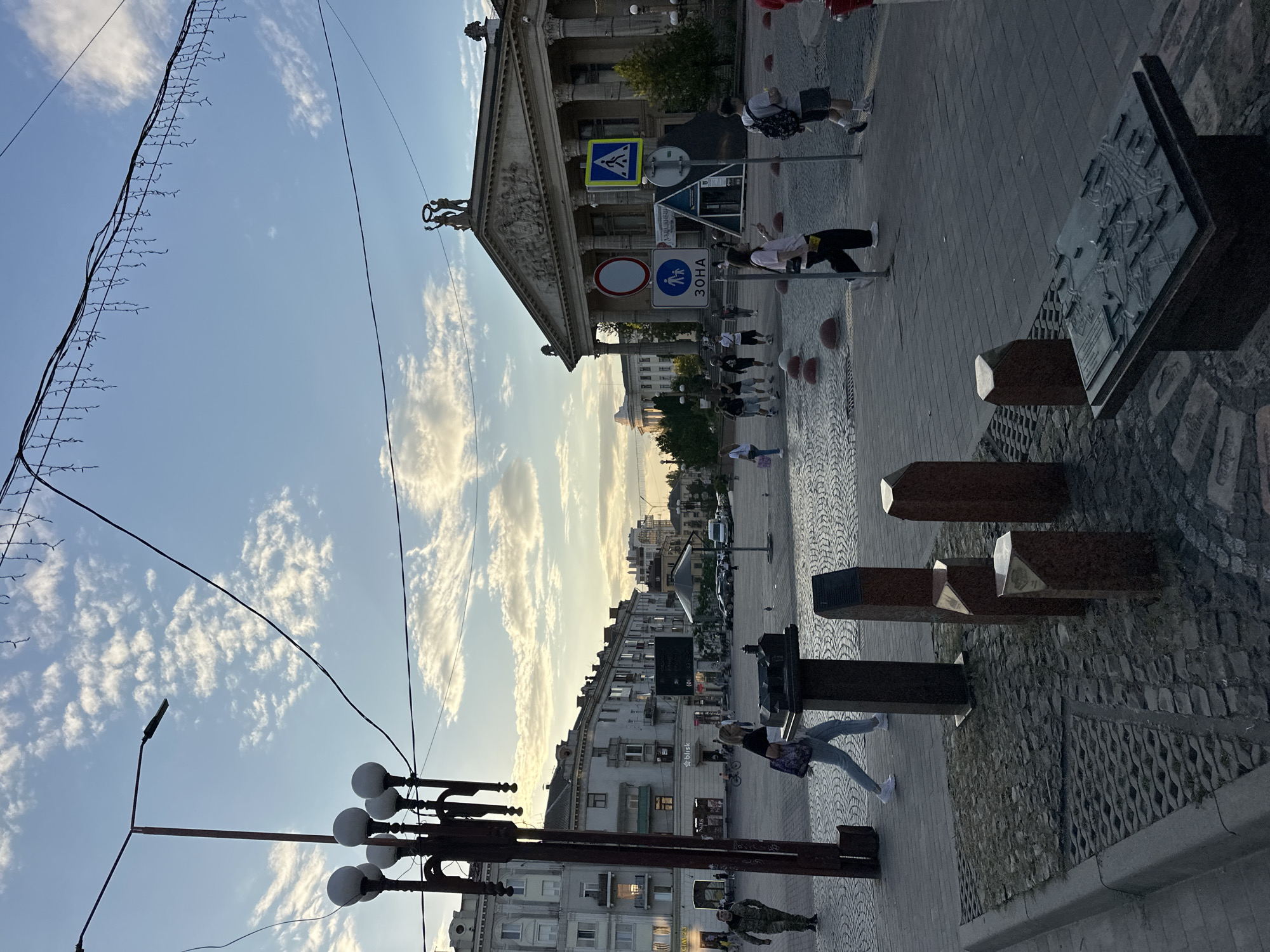 The image captures a scene in Ternopil, Ukraine, likely at Theater Square. The photograph is taken at dusk, with a warm, golden light illuminating the sky and casting long shadows. A grand neoclassical building, presumed to be the Ternopil Academic Regional Ukrainian Music and Drama Theater, dominates the background with its imposing columns and sculptural details. In the foreground, a paved plaza with intricate cobblestone patterns is visible. Several people are walking across the plaza, some individually, others in small groups, suggesting a relaxed evening atmosphere.
On the left side of the frame, a prominent lamppost with multiple lights stands tall, a common feature in urban streetscapes. A soldier in camouflage uniform is seen walking near the lamppost. Several smaller, reddish-brown granite monuments or markers are placed in the foreground, adding to the plaza's aesthetic. A detailed, three-dimensional model of a city or historical site rests on a pedestal in the lower right corner. Traffic signs, including a pedestrian crossing sign with a blue background and a white silhouette of a person, are visible on the right. The sky is a pale blue, dotted with wispy clouds that catch the fading sunlight. Decorative lights, possibly for an event or holiday, are strung across the upper portion of the image, adding a festive touch. The overall impression is one of a calm, urban evening with a blend of architectural grandeur and everyday life.