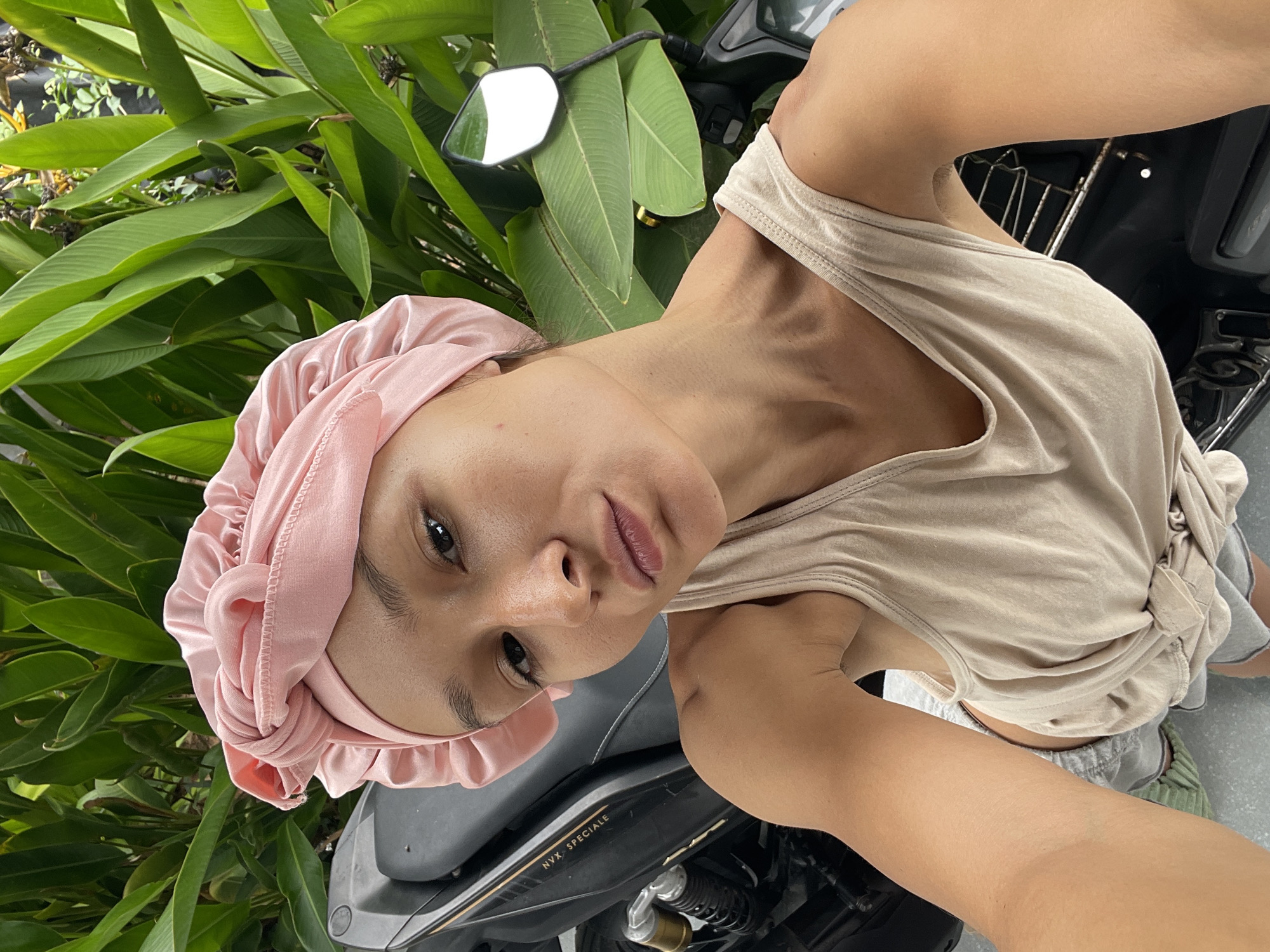 A woman wearing a pink head covering and a beige tank top is posing for a selfie while sitting on a black motorcycle. Green plants are visible in the background. The motorcycle has the words "NVX SPECIALE" written on it. The location is Phu Thuy Ward, Vietnam.