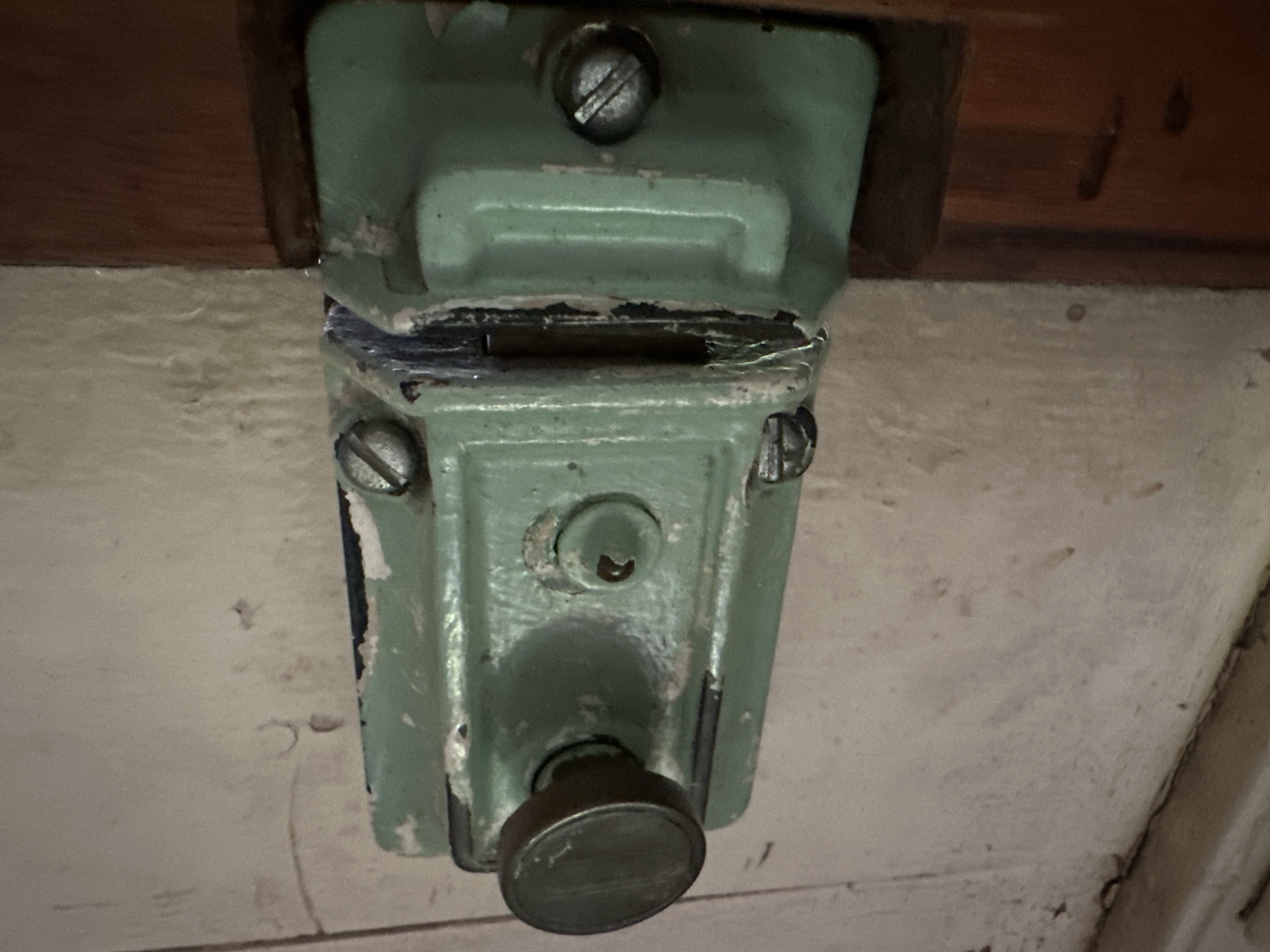 The image shows a close-up, horizontal view of a weathered, light green metal door latch mechanism attached to a wooden door frame. The latch itself is rectangular with a rotating knob on the left side, which is made of a darker metal. There are visible screw heads securing the latch to the frame. The paint on the latch is chipped and worn, revealing darker layers underneath and some rust. To the right of the latch, a strike plate is mounted on the door frame. The surrounding wall appears to be painted a light, off-white color and shows signs of age and wear, including scuff marks and possibly water stains. The lighting is uneven, casting shadows on parts of the latch and frame. There is no indication of people or any specific location in Thora, Australia, present in the image.