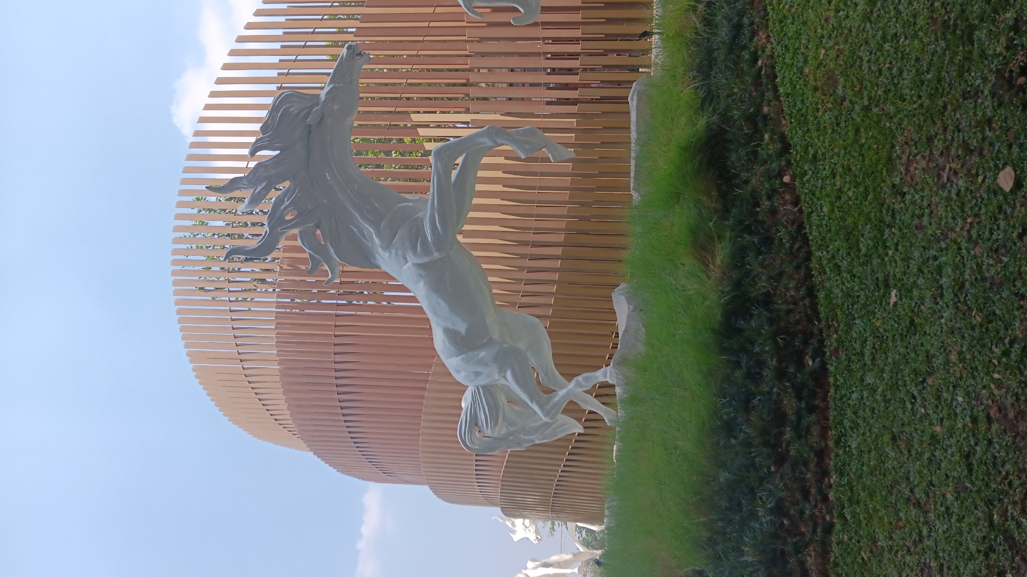 A white horse sculpture is prominently displayed in front of a modern architectural structure in Bekasi, Indonesia. The sculpture depicts the horse in mid-leap, with its mane and tail flowing dynamically. The architectural element behind the sculpture is a curved facade made of vertical wooden slats, creating a cylindrical or spiral effect. The scene is set outdoors, with lush green grass in the foreground and a bright, clear blue sky above, with some scattered clouds. There are other horse sculptures visible in the background, suggesting this is a display or park area. The lighting suggests it is daytime.