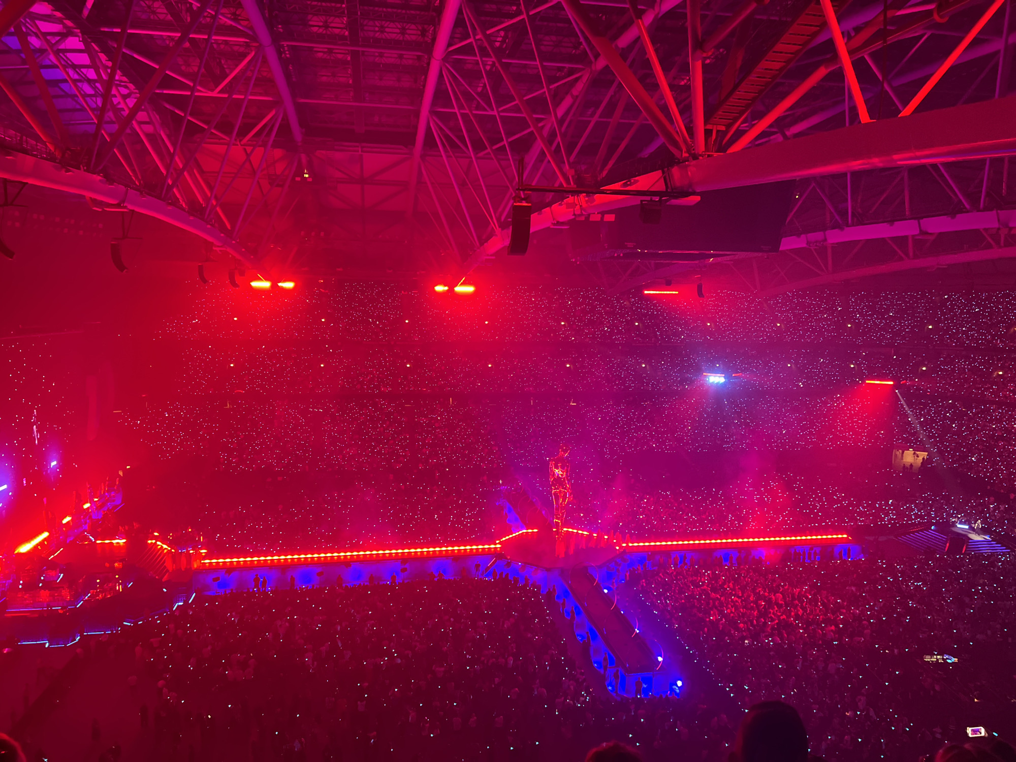 The image depicts an aerial view of a concert taking place in the Friends Arena in Solna, Sweden. The arena is filled with a vast crowd, each person holding up a light, creating a sea of small, white dots against the predominantly red and pink lighting. The stage is illuminated, and a performer is visible standing on a raised platform. The arena's architectural structure, with its exposed beams and lighting rigs, is prominent in the upper portion of the image. The overall atmosphere is that of a large-scale live music event.