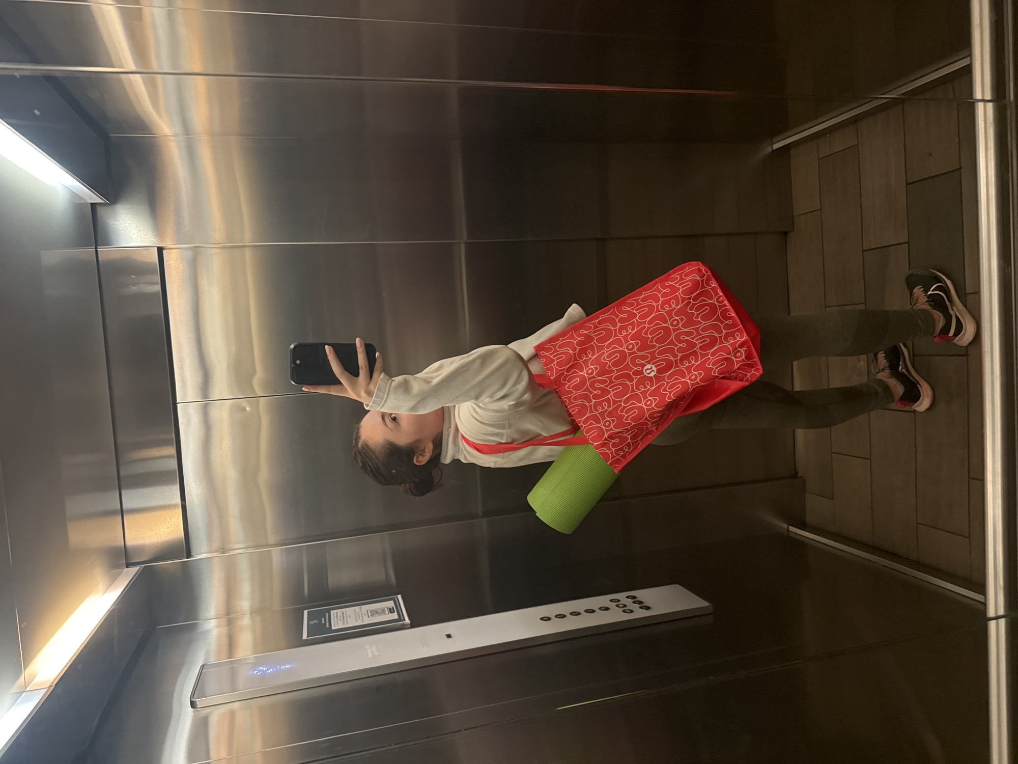 A woman is taking a selfie in an elevator. She is wearing a white sweatshirt, olive-green leggings, and black and white running shoes. She has a red tote bag with white lettering and a green yoga mat. The elevator has stainless steel walls and a control panel with buttons. The floor of the elevator appears to be made of dark tiles. Based on the design of the elevator control panel, it could possibly be located in Mexico City.