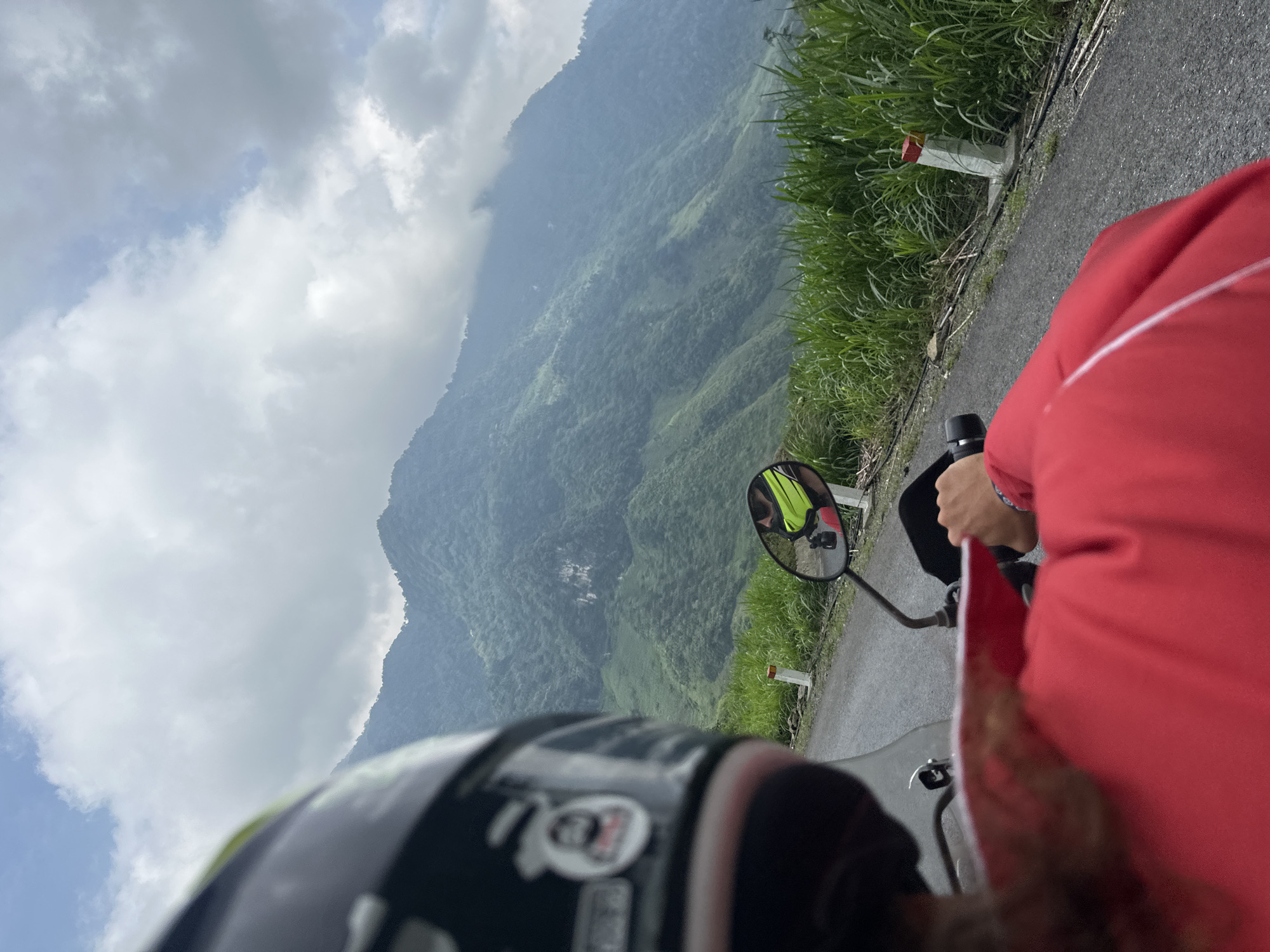 The image captures a first-person perspective from a motorbike, showcasing a scenic ride through a mountainous landscape in Minh Sơn Commune, Vietnam.
In the foreground, the back of a black and white motorcycle helmet is visible on the left, featuring a sticker that resembles the "AGV" logo. To the right, the arm and torso of a person wearing a bright red jacket are seen, likely a passenger. The motorbike's handlebars and a rearview mirror are also prominent. The mirror reflects another individual, presumably the driver, wearing a bright yellow or lime green reflective vest.
The motorbike is on an asphalt road that curves alongside the mountainous terrain. Lush green grass and dense foliage line the roadside, and a concrete road marker with a distinctive red and white painted top is visible. Beyond the road, vast, verdant mountains covered in thick forests stretch into the distance, partially obscured by mist or clouds, indicating a high altitude and potentially humid environment. The sky is partly cloudy with some clear blue patches, suggesting it is daytime. The scene conveys a sense of adventure and travel through a beautiful natural setting.