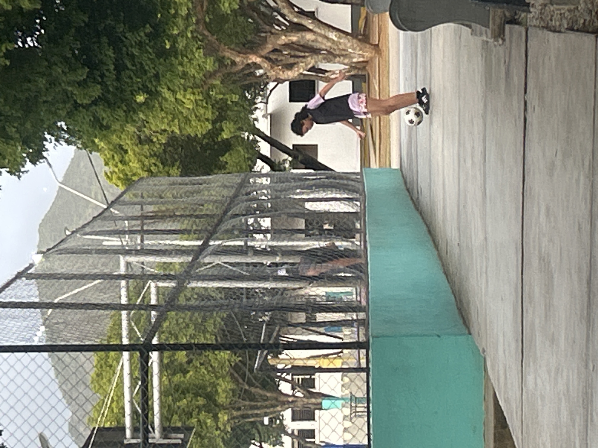 A young person, with dark hair pulled back, stands on a concrete walkway in what appears to be an outdoor recreational area, possibly a school or park, in Santa Ana, Venezuela. They are wearing a dark sleeveless vest over a light pink shirt and patterned shorts, with black athletic shoes. The person is in the process of kicking or controlling a soccer ball with their right foot. Behind them, a chain-link fence encloses a sports court, with a turquoise-colored wall visible along the edge of the walkway. In the background, there are trees with lush green foliage and a building with white walls and dark windows. Beyond the trees and buildings, a large, verdant mountain range dominates the skyline under a hazy sky.