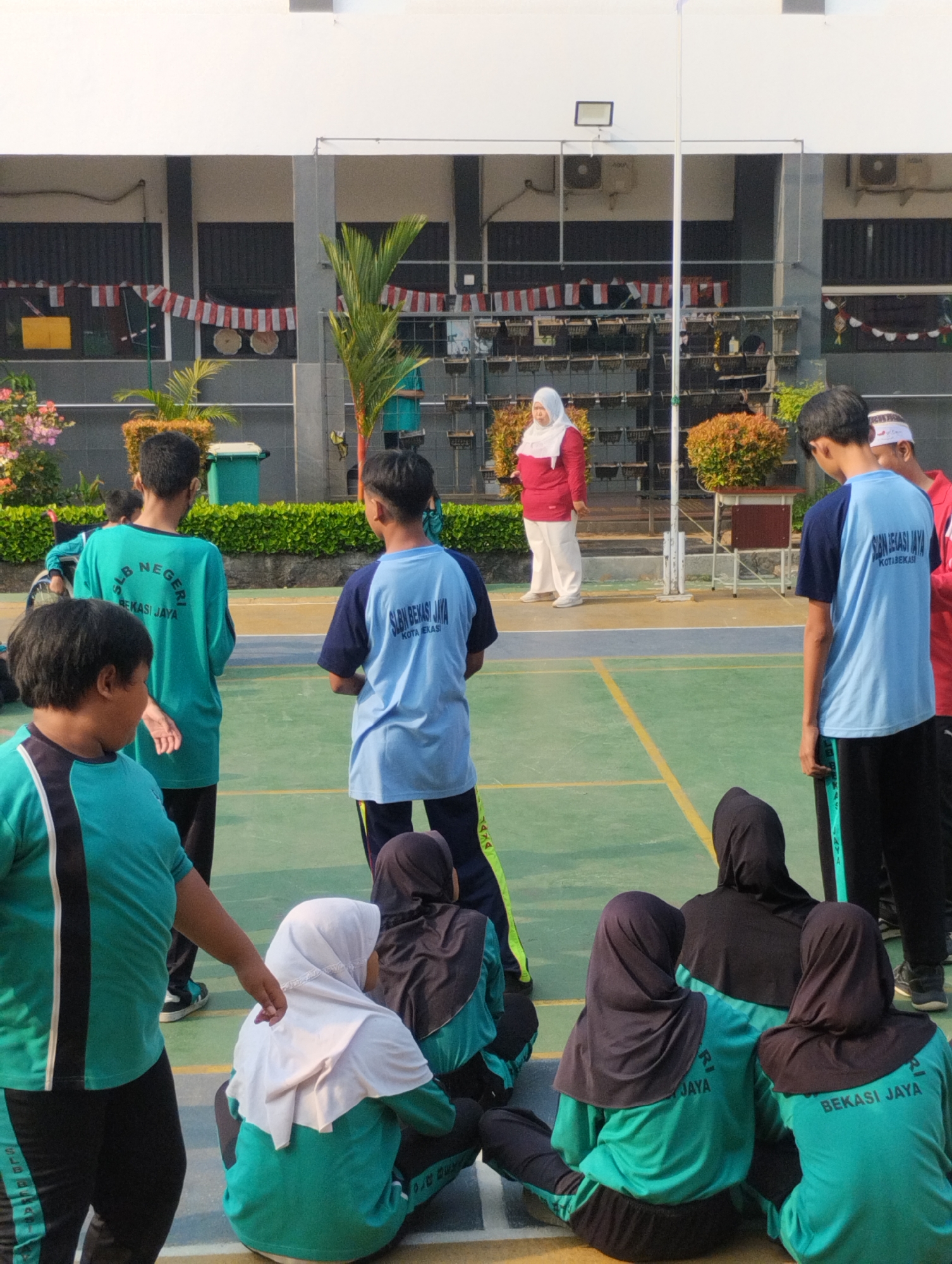 This is a medium shot, eye-level photograph taken outdoors on a sunny day. The scene appears to be a schoolyard in Bekasi, Indonesia, likely during an outdoor activity or assembly. In the foreground, several young people are visible, some sitting and others standing. The majority are wearing teal and black athletic uniforms with the inscription "SLB NEGERI BEKASI JAYA" and "KOTA BEKASI" on their backs, suggesting they are students from the State Special School of Bekasi.
Several individuals are wearing hijabs, indicating a Muslim population. One young person in the foreground, facing away from the camera, is wearing a teal shirt and black pants. To their left, another individual in a similar teal uniform is partially visible.
In the midground, a group of students in teal uniforms are standing, facing away from the camera. To the right, two individuals in light blue uniforms with "SLBN BEKASI JAYA" and "KOTA BEKASI" visible on their shirts are standing. A woman in a red long-sleeved shirt and white pants, wearing a white hijab, stands near a flagpole in the background, appearing to address the students.
The environment consists of a green court with yellow lines, characteristic of a sports court. Behind the students, there is a building with grey walls and windows, decorated with red and white Indonesian flags or banners. Some potted plants and trees are also present in the scene, and a trash bin is visible near the building. The overall atmosphere suggests an organized school event in an urban setting.
