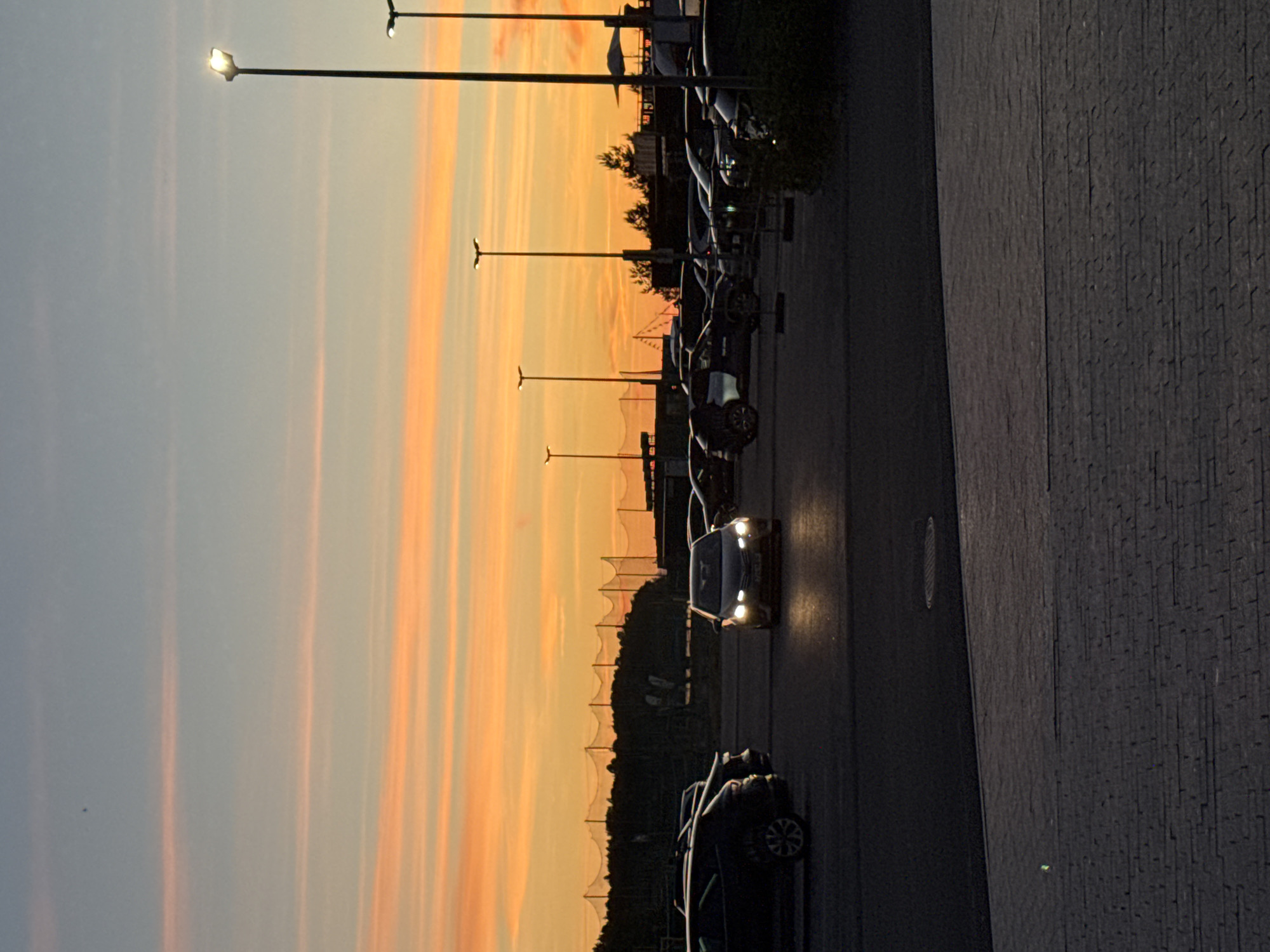 An outdoor, eye-level, medium shot captures a parking lot scene during sunset, with a vibrant orange and yellow sky streaked with thin, pale pink clouds. In the foreground, a paved area transitions to asphalt where several cars are parked. A dark-colored sedan is positioned in the center, its headlights illuminated and casting a glow on the wet asphalt. Other vehicles, mostly dark SUVs and sedans, are visible throughout the lot. Streetlights, tall and slender, are scattered across the scene, with one in the mid-right illuminated, its light source a bright white. Behind the parking lot, silhouetted trees and a fence with netting, possibly for a sports field, are visible against the colorful sky. The overall impression is of a quiet evening in a parking area, enhanced by the dramatic sunset. This image was taken in Tallinn, Estonia, near the Pirita beach area, specifically in the parking lot of the Pirita marina.