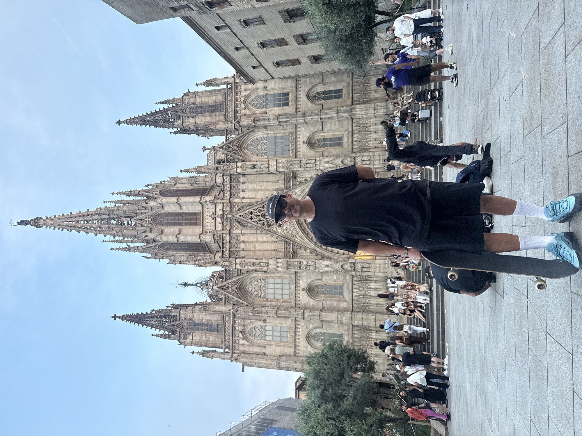 A full-length portrait shows a man standing with a skateboard in front of the Barcelona Cathedral. The man is wearing a black t-shirt, black shorts, white socks, and blue athletic shoes. He also has a baseball cap on and sunglasses. He is holding the skateboard in front of him and looking slightly to the right. In the background is the gothic cathedral. The square in front of the cathedral is filled with tourists who are sitting on the steps or standing and looking at the building. The sky is blue.