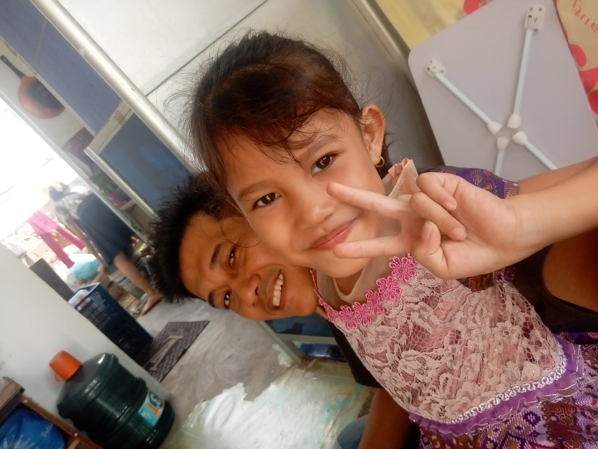 In Batam, Indonesia, two individuals, a child and an adult male, are pictured in what appears to be an indoor setting, possibly a home or a communal area. The child, positioned in the foreground, is looking towards the camera and making a peace sign with her right hand. She is wearing a dress adorned with intricate floral lace. The adult male is visible behind and to the side of the child, also facing the camera, with a clear smile. Behind them, a large green water jug is situated, suggesting a common household appliance. The overall scene is captured in a casual, selfie-style photograph, with the background slightly blurred, indicating a close-up shot.