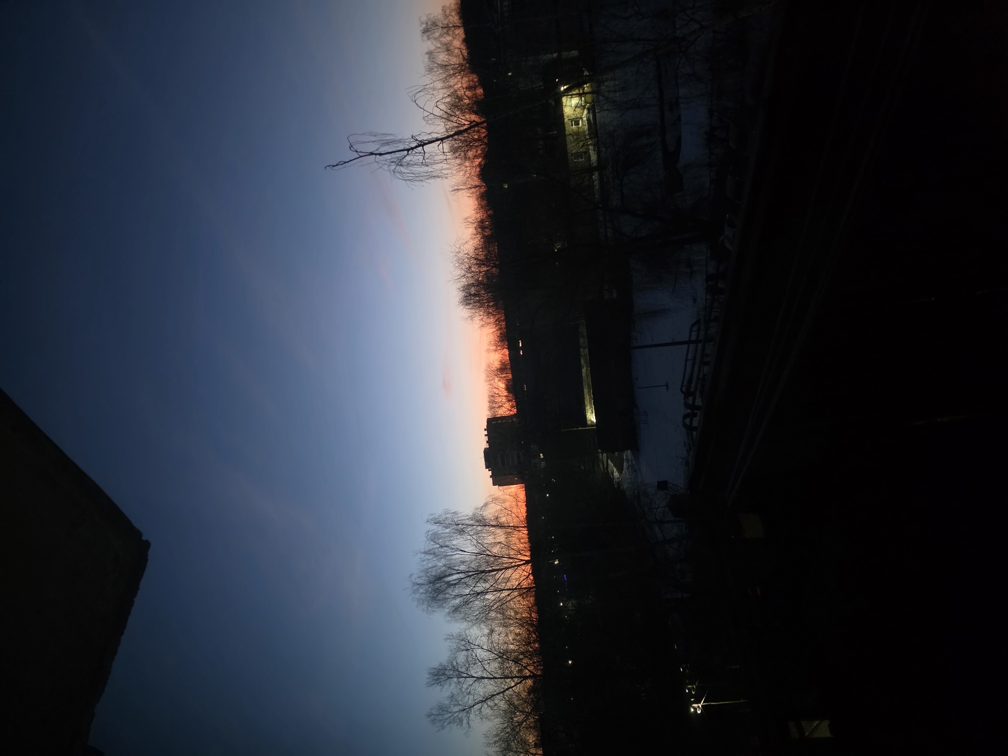 The image depicts a sunset in Chaikovsky, Russia, with a gradient sky ranging from dark blue to orange and red near the horizon. Silhouetted trees and buildings line the horizon. Streetlights are visible. Snow is on the ground in some areas. The foreground is dark, suggesting a view from a window or balcony.