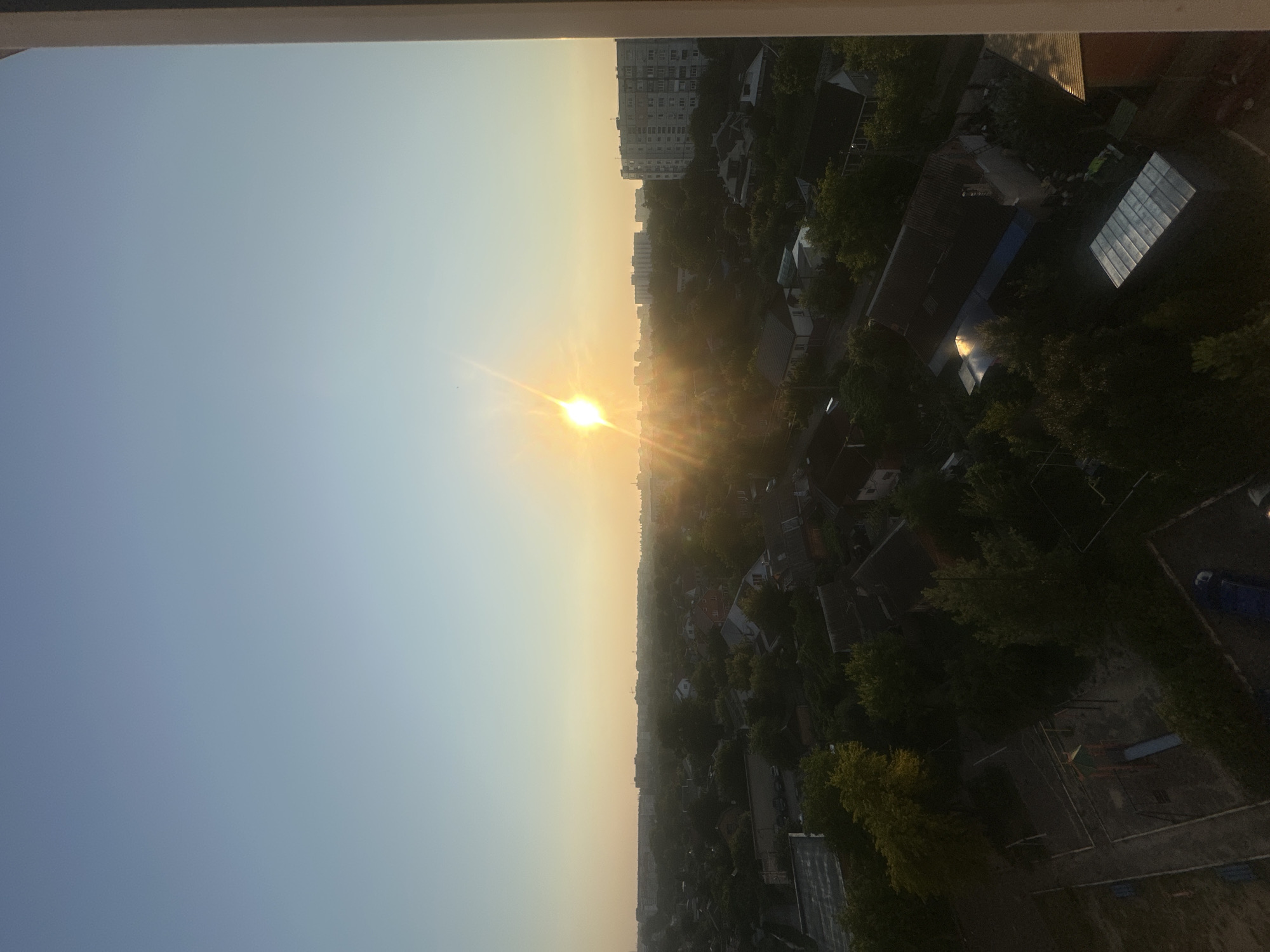 This is a high-angle, wide shot of a residential area at sunrise or sunset. The sun is low on the horizon, casting a warm glow and creating lens flare. The sky is a clear, pale blue that transitions to a soft yellow near the horizon. In the foreground, on the right side of the frame, a white vertical structure, possibly a building edge or balcony railing, partially obscures the view.
The middle ground is filled with a dense canopy of green trees interspersed with the roofs of houses. Many of the houses appear to be single-family dwellings with red-tiled or dark-colored roofs. Some houses are partially obscured by the foliage. In the distance, several multi-story apartment buildings are visible, suggesting a mix of suburban and urban development. The buildings are gray and have a blocky, utilitarian architectural style. A few faint, skeletal structures that might be construction cranes or communication towers are also present on the distant skyline.
In the lower left portion of the image, a small playground with a slide and climbing structure is visible. A paved pathway or courtyard surrounds the playground. Darker shadows suggest the early morning or late evening light. The overall impression is one of a quiet, residential neighborhood at the beginning or end of the day.