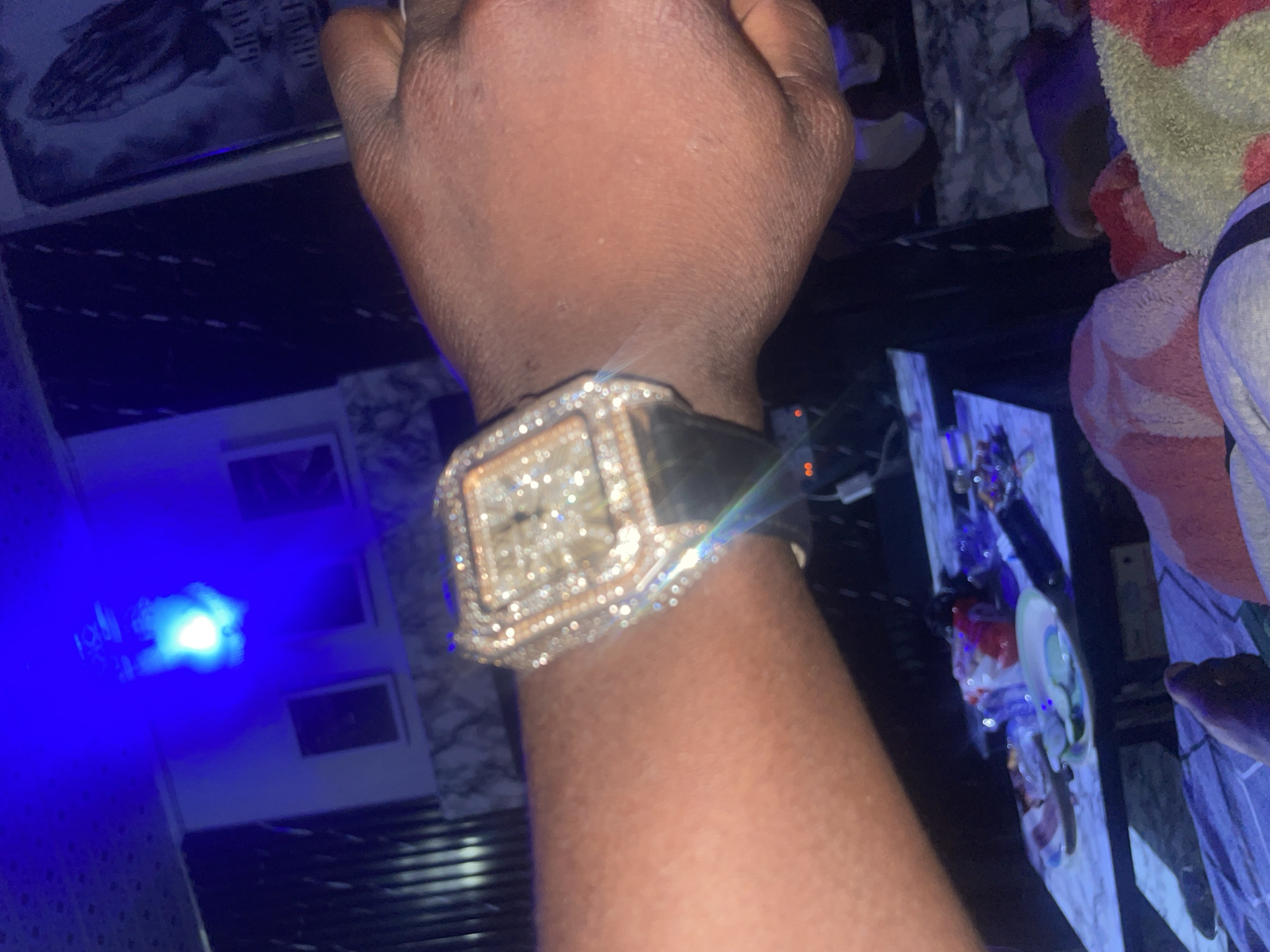 The image shows a close-up of a person's wrist wearing a diamond-encrusted watch. The watch face is square with a black band. The background is dark and appears to be an indoor setting with blue lighting. There are several framed pictures on the wall, one of which depicts praying hands. A table with some objects, possibly food or drinks, is visible in the lower part of the image. The overall impression is one of luxury and a nighttime atmosphere. The specific location in Rafin Par, Nigeria cannot be determined from the image.
