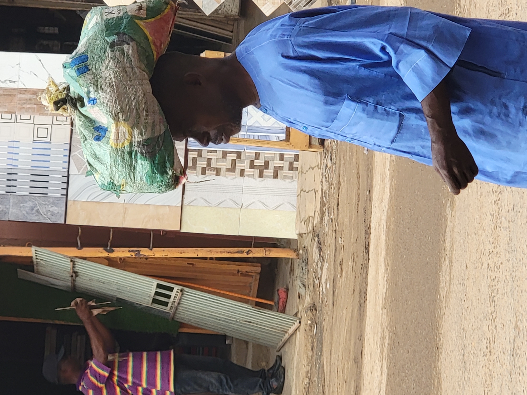 A man in a blue shirt carries a large, heavy bag on his head in Maiburiji, Nigeria. He is standing near a display of tiles. Another man in a purple and yellow striped shirt is seen in the lower part of the frame, appearing to be cleaning or inspecting something on a rack. The environment seems to be a roadside or market area, with a sandy or dirt-like surface.