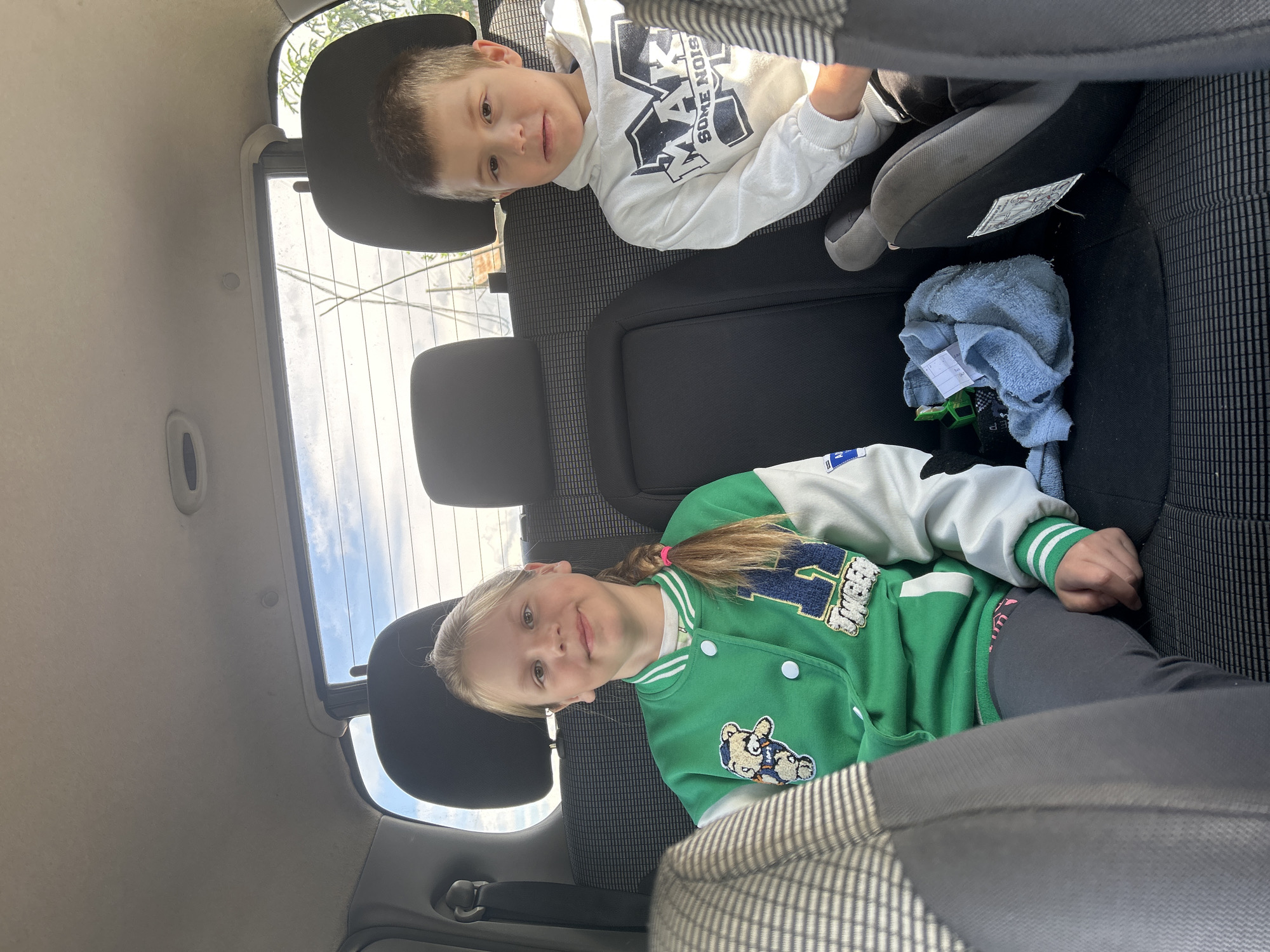 Two children are sitting in the back of a car. A boy in a white hoodie is in the upper portion of the frame, and a girl in a green jacket is in the lower portion. A car seat is also visible. The background shows a car interior with headrests and a view of the sky through the back window. The location is not identifiable as Фоки, Russia from the given image.