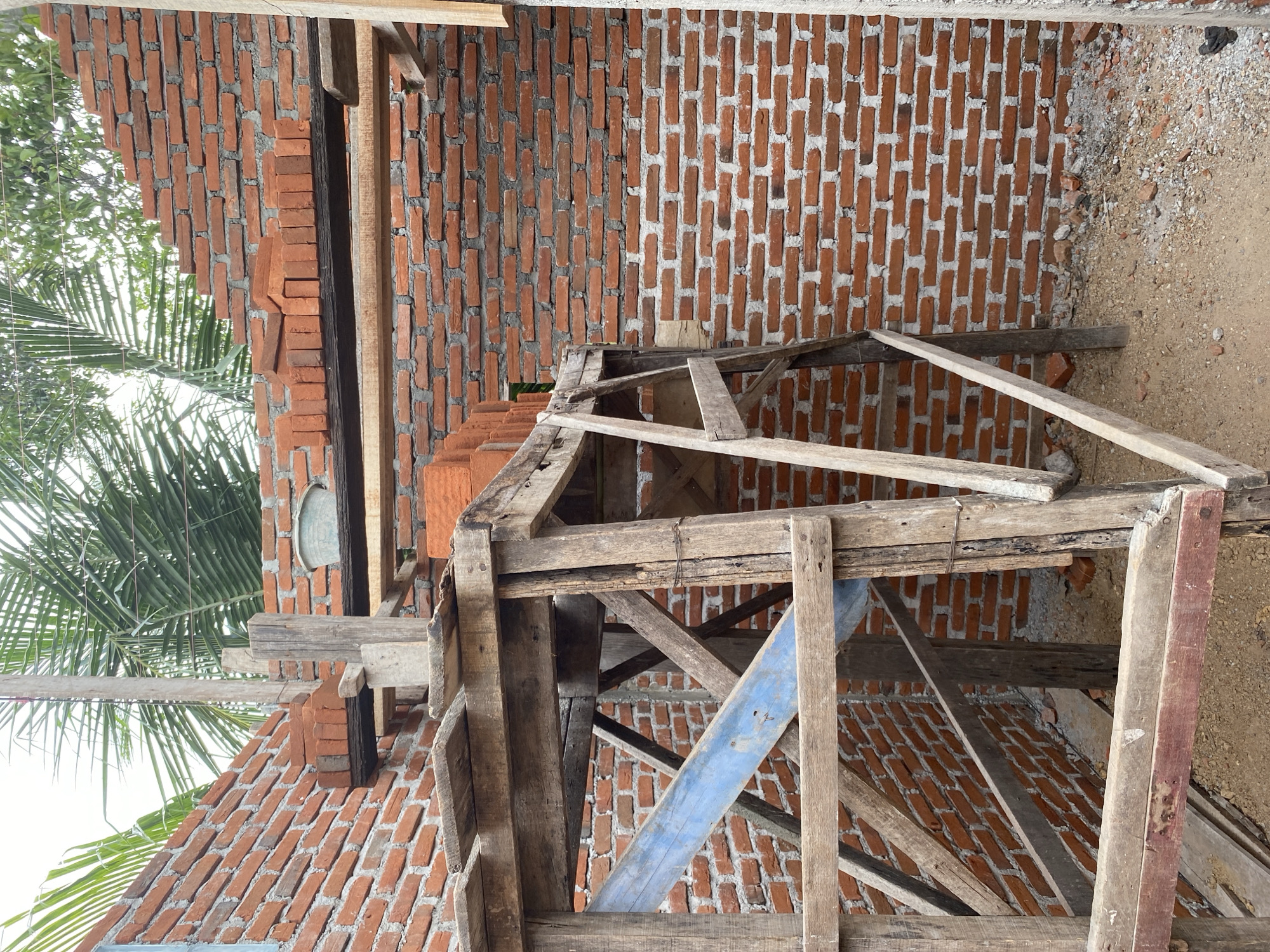 A construction site shows brick walls being built. Wooden scaffolding is erected on the left, supporting planks of wood and a blue plank. Stacked bricks are visible on the scaffolding. Palm fronds and greenery are seen in the background, suggesting an outdoor, possibly tropical, location. The ground is covered in sand and debris.