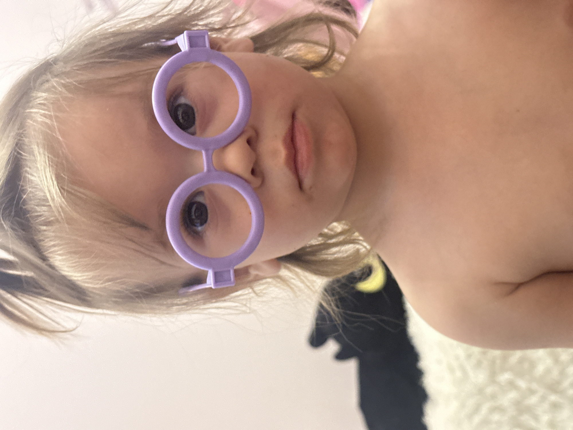 A fair-skinned child with blonde hair wears purple, round toy glasses. The child's chest and shoulders are bare. A black and white stuffed animal is partially visible behind the child. The background appears to be a light-colored wall and bed covering. The image is rotated 90 degrees. There is no indication of Kharnavy, Russia.