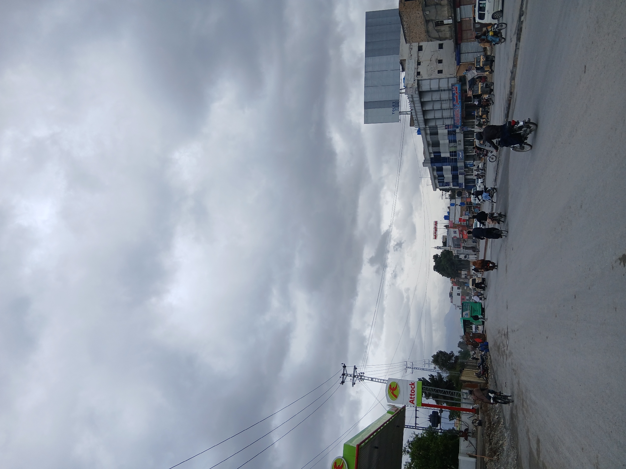 The image shows a street scene in Quetta City Tehsil, Pakistan, under a cloudy sky. A petrol station with an "Attock" logo is visible on the lower left. The street is lined with buildings of varying heights, and numerous parked and moving motorcycles are present, suggesting active traffic. Several individuals are seen riding motorcycles along the street. Power lines and utility poles are also visible, adding to the urban environment.