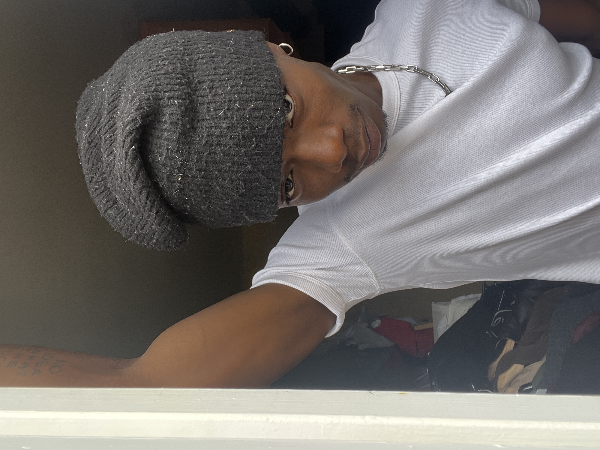 A person wearing a gray beanie, silver hoop earring, white t-shirt, and silver chain is partially visible. The person is leaning over a white surface, possibly a window sill, with their right arm resting on it. The person's left arm is also visible, adorned with tattooed writing. The background is a dark, indistinct space with clothes and other objects visible underneath. There is no information to indicate this picture was taken in Barcelona, Spain.