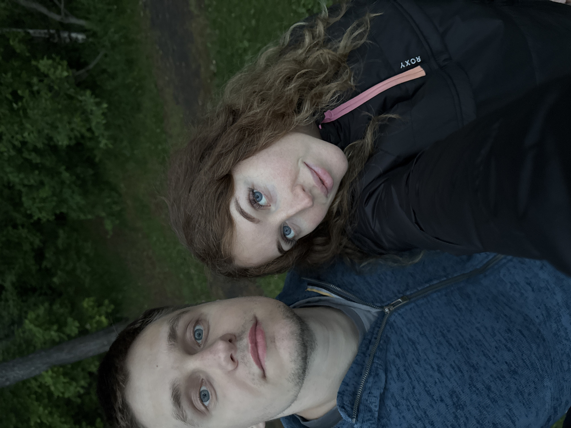 The image shows a selfie of a man and a woman. The woman has curly brown hair and blue eyes. She is wearing a black jacket with a pink accent. The man has short brown hair and blue eyes. He is wearing a blue jacket with a gray shirt underneath. The background appears to be a wooded area. The location is in the city of Chaikovsky, Russia.