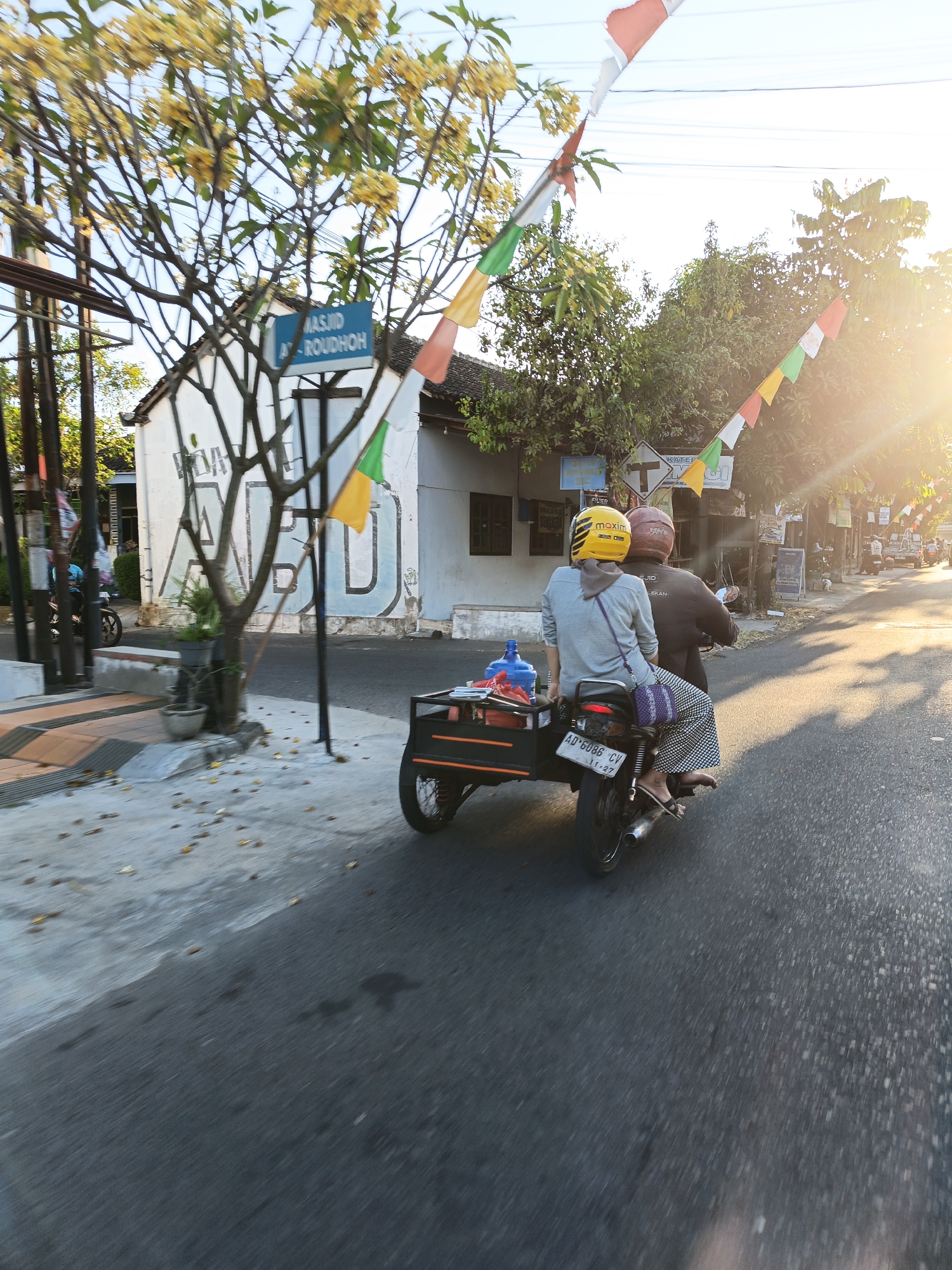 In the city of Bareng Lor, Indonesia, a snapshot captures a scene on a sun-drenched road. Two individuals are seen riding a motorcycle, the passenger in front wearing a yellow helmet with "maxim" branding, and the person behind wearing a red helmet. They are traveling alongside a small trailer attached to the motorcycle. In the background, a white building with a blue sign that reads "MASJID AL-ROUDOH" is visible, along with trees and colorful decorative flags strung across the scene. The road is paved, and scattered leaves suggest an outdoor environment. The overall lighting indicates it is likely late afternoon.