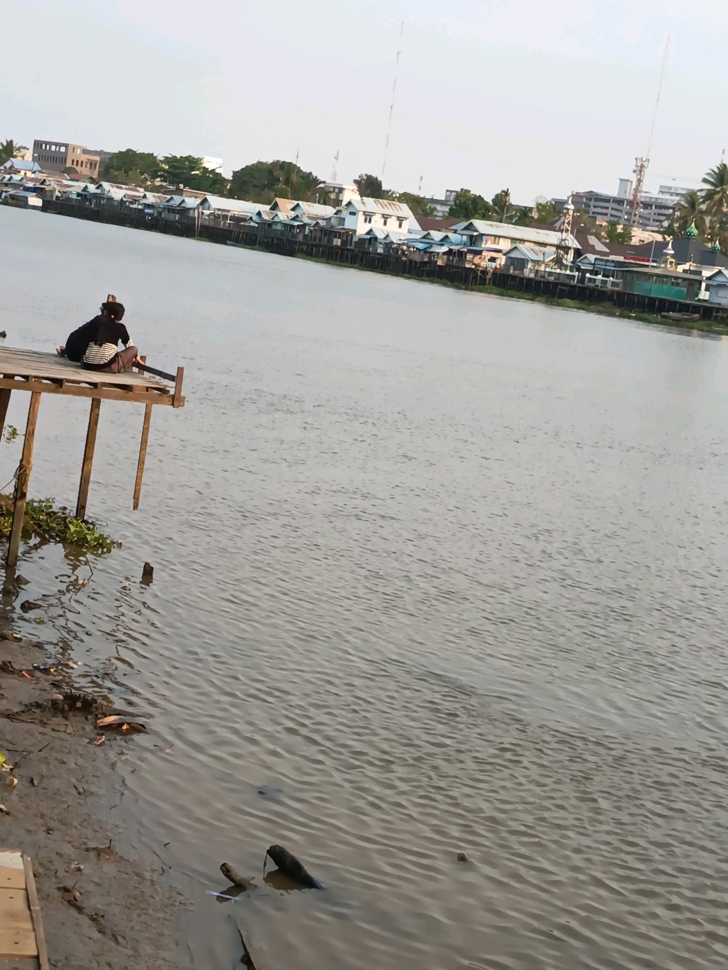 The image depicts a wide river in Banjarmasin, Indonesia, with a stilted settlement along the far bank. The settlement consists of numerous small houses with blue and white exteriors and corrugated metal roofs, built on wooden stilts over the water. In the foreground, two individuals, possibly a parent and child, sit on a wooden pier that extends into the river. The person in the back wears a black garment and the person in front wears a striped shirt and dark pants. The river is calm with ripples on the surface. The muddy riverbank on the left side of the frame is littered with debris. The sky is overcast, suggesting either early morning or late afternoon light.