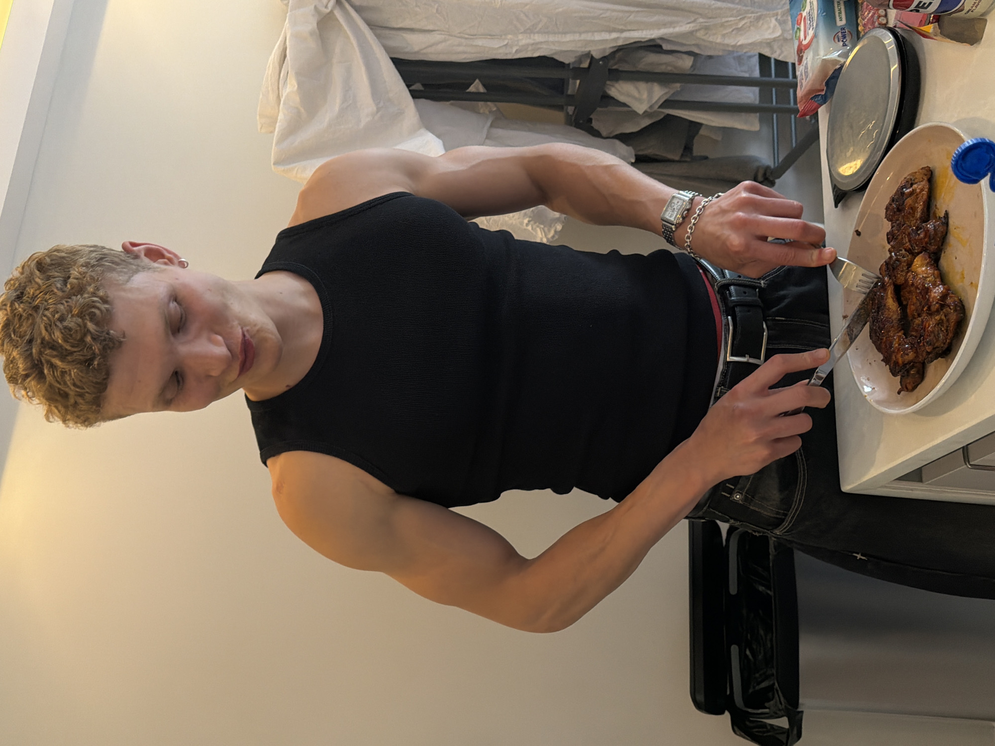 A young man with curly blonde hair and a black tank top is standing in a kitchen, cutting meat on a plate with a knife and fork. He is wearing dark jeans and a watch on his left wrist. The kitchen counter is white and has a trash can and other items on it. The background shows a wall and some draped fabric. The image was taken in London, United Kingdom.